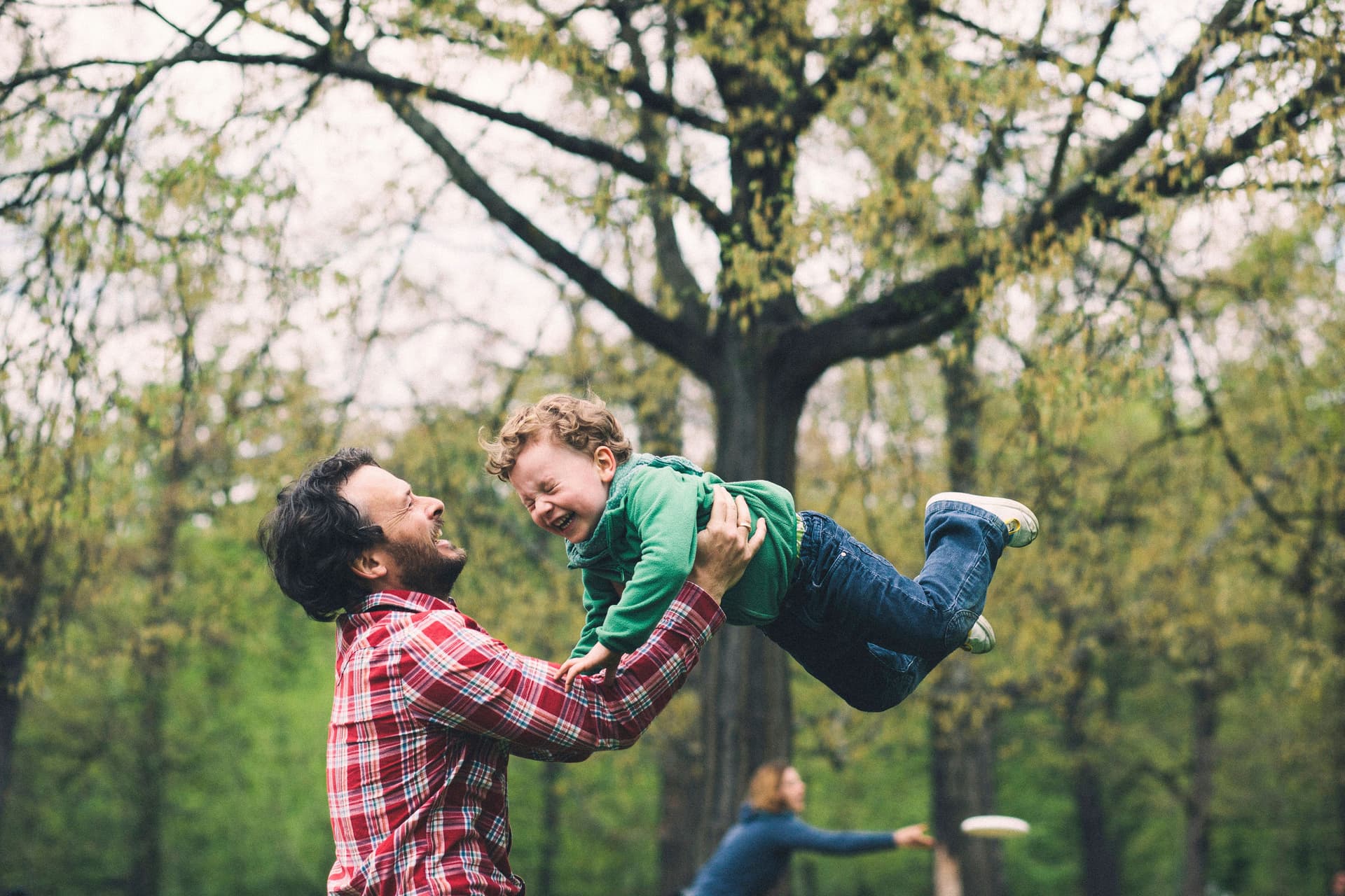 Vater wirbelt seinen Sohn im Park herum. Die Mutter spielt im Hintergrund Frisbee.