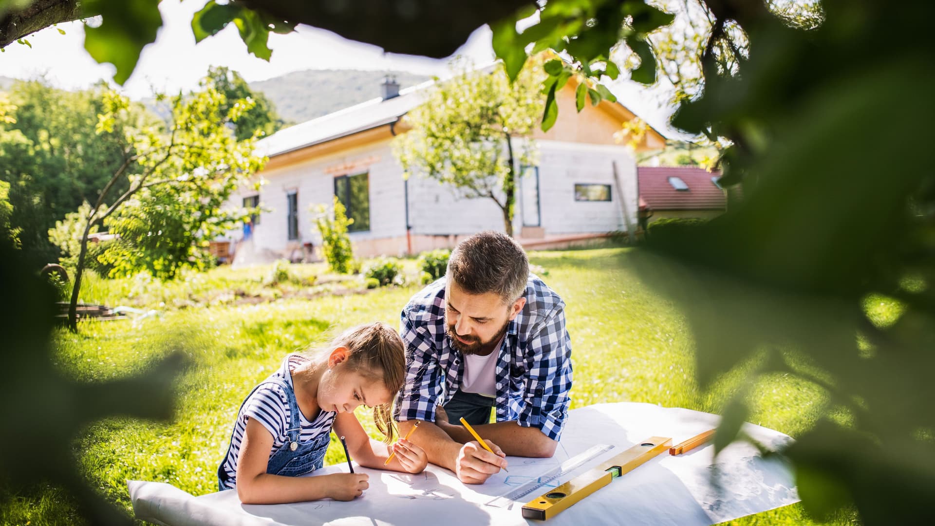 Vater und Tochter planen etwas im Garten mit Stift und Papier. Im Hintergrund steht ein Haus und im Vordergrund ein Baum.