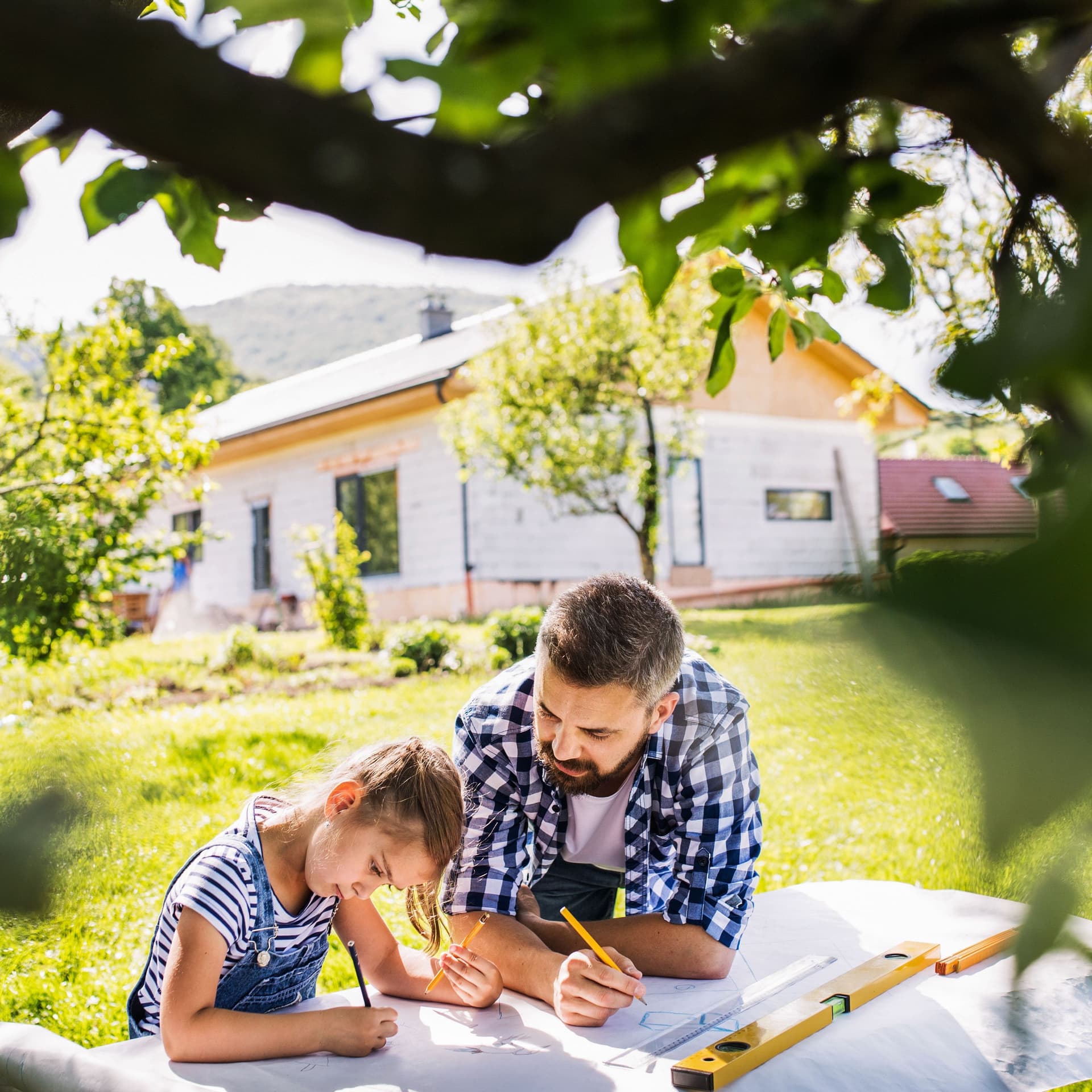 Vater und Tochter planen etwas im Garten mit Stift und Papier. Im Hintergrund steht ein Haus und im Vordergrund ein Baum.