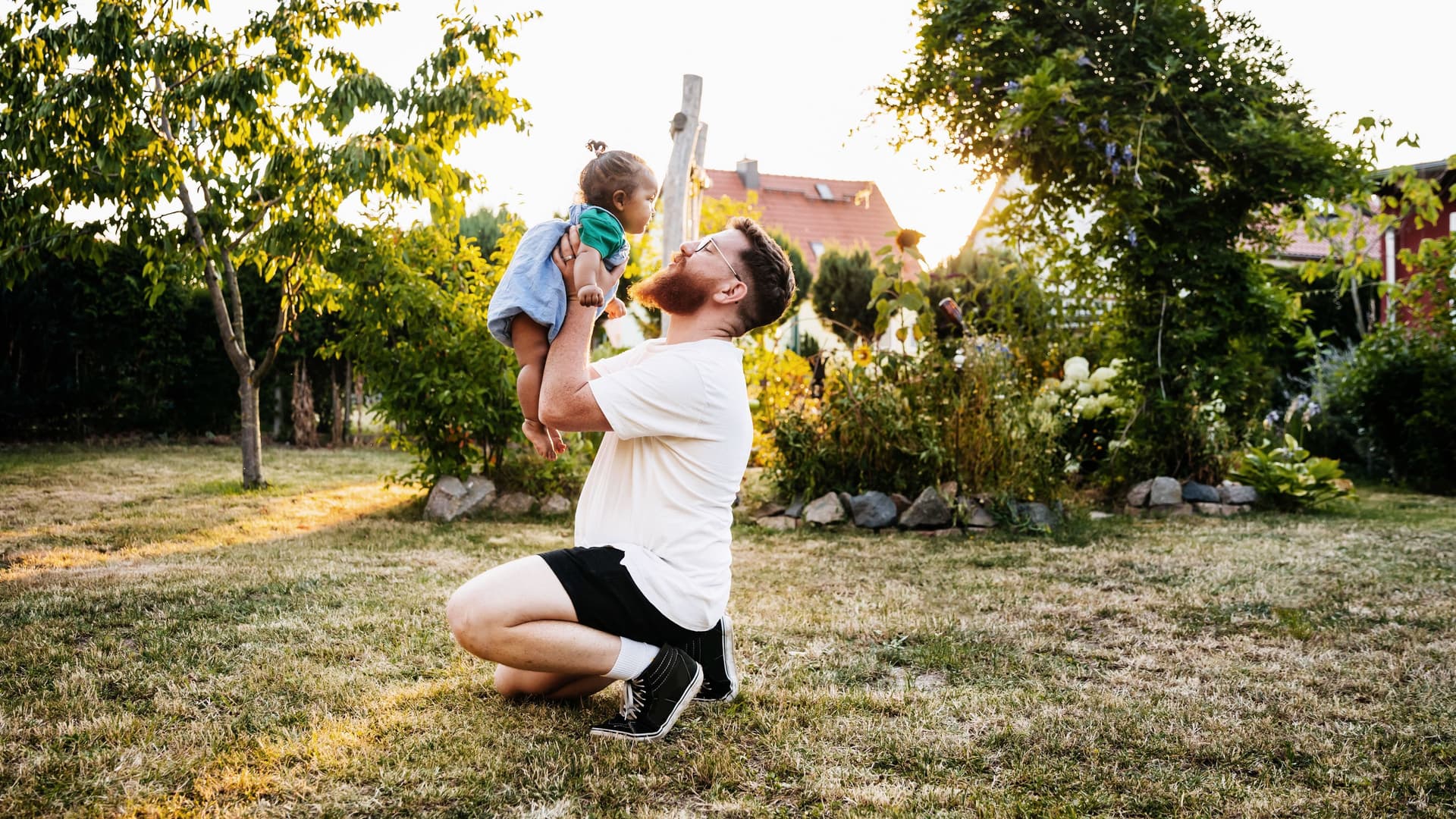 Vater kniet auf dem Rasen und hebt seine kleine Tochter im Garten vor sich hoch. 