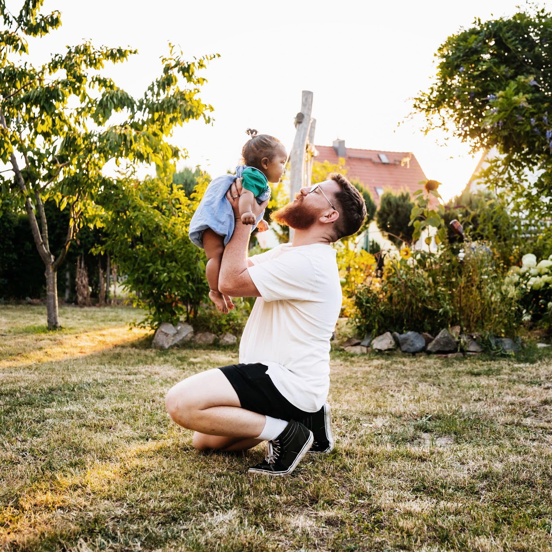 Vater kniet auf dem Rasen und hebt seine kleine Tochter im Garten vor sich hoch. 