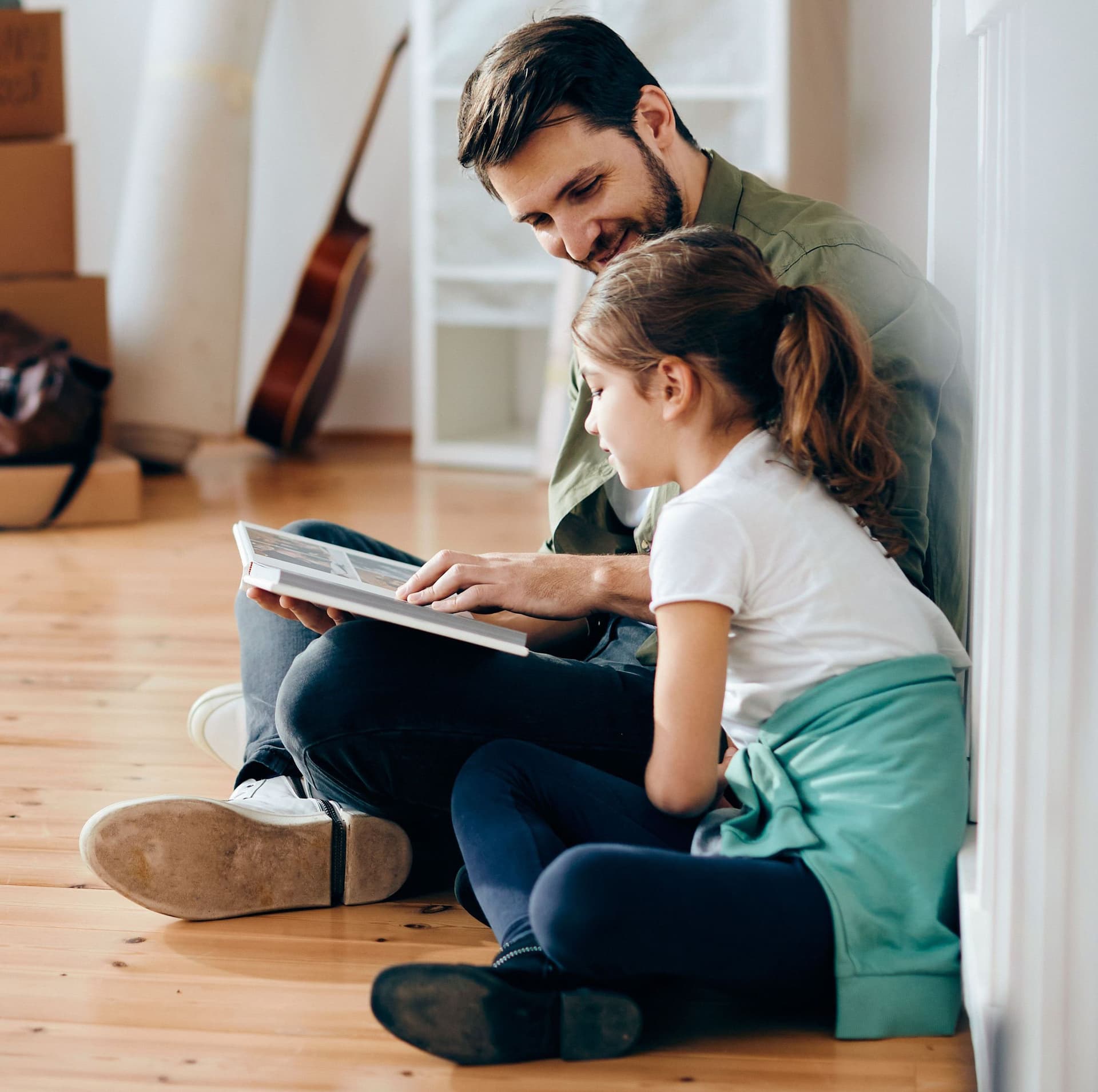 Lächelnder Vater und kleine Tochter sitzen auf dem Holzboden und gucken ein Fotoalbum an. Der Raum ist fast leer und im Hintergrund sind Umzugskisten.
