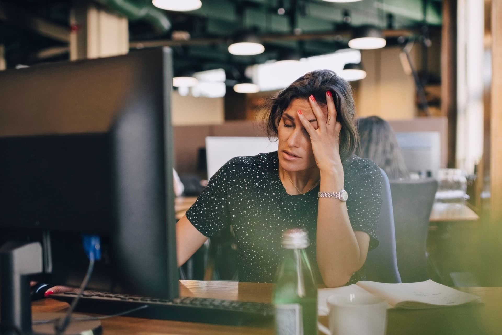 Eine Frau sitzt an einem Schreibtisch in einem Büro vor einem Computer und hält ihre Hand an den Kopf. Sie hat die Augen geschlossen und wirkt erschöpft.