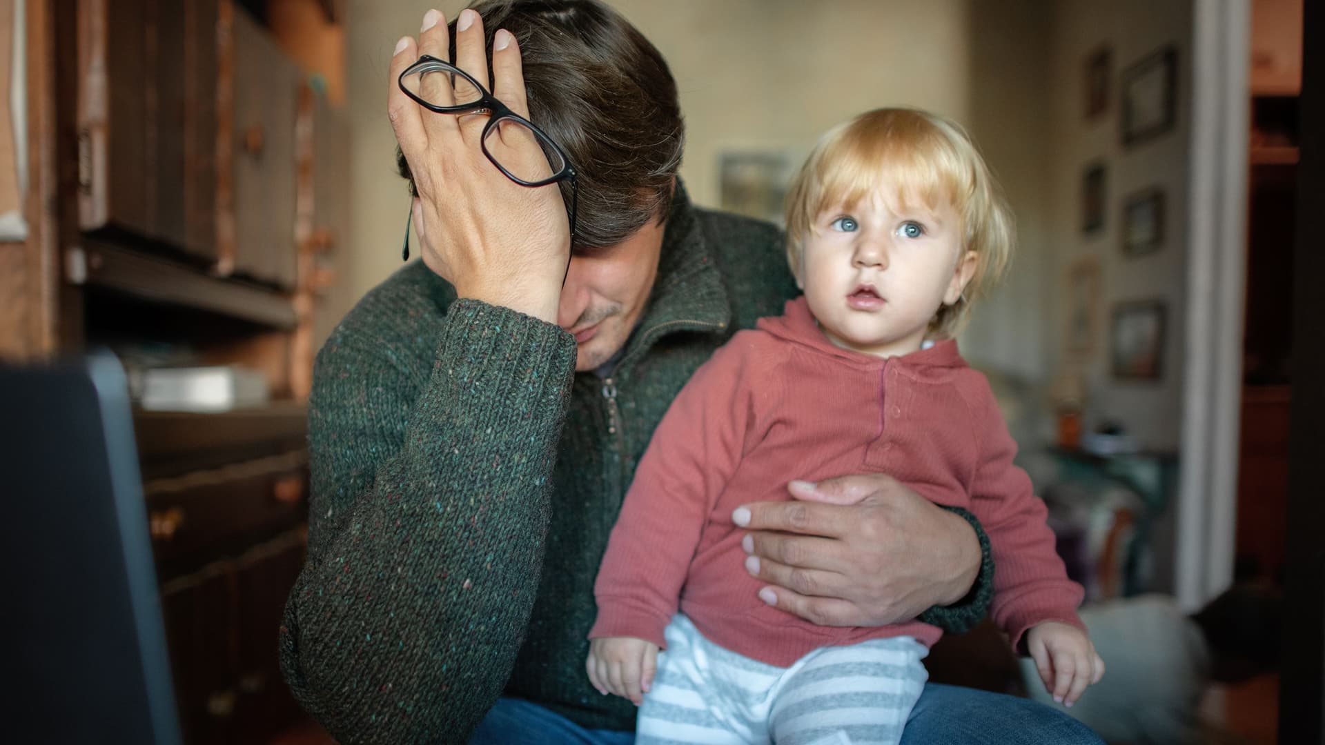Ein Mann hält sich verzweifelt das Gesicht. Auf seinem Schoss sitzt ein Kleinkind. Auf dem Tisch liegen Rechnungen.