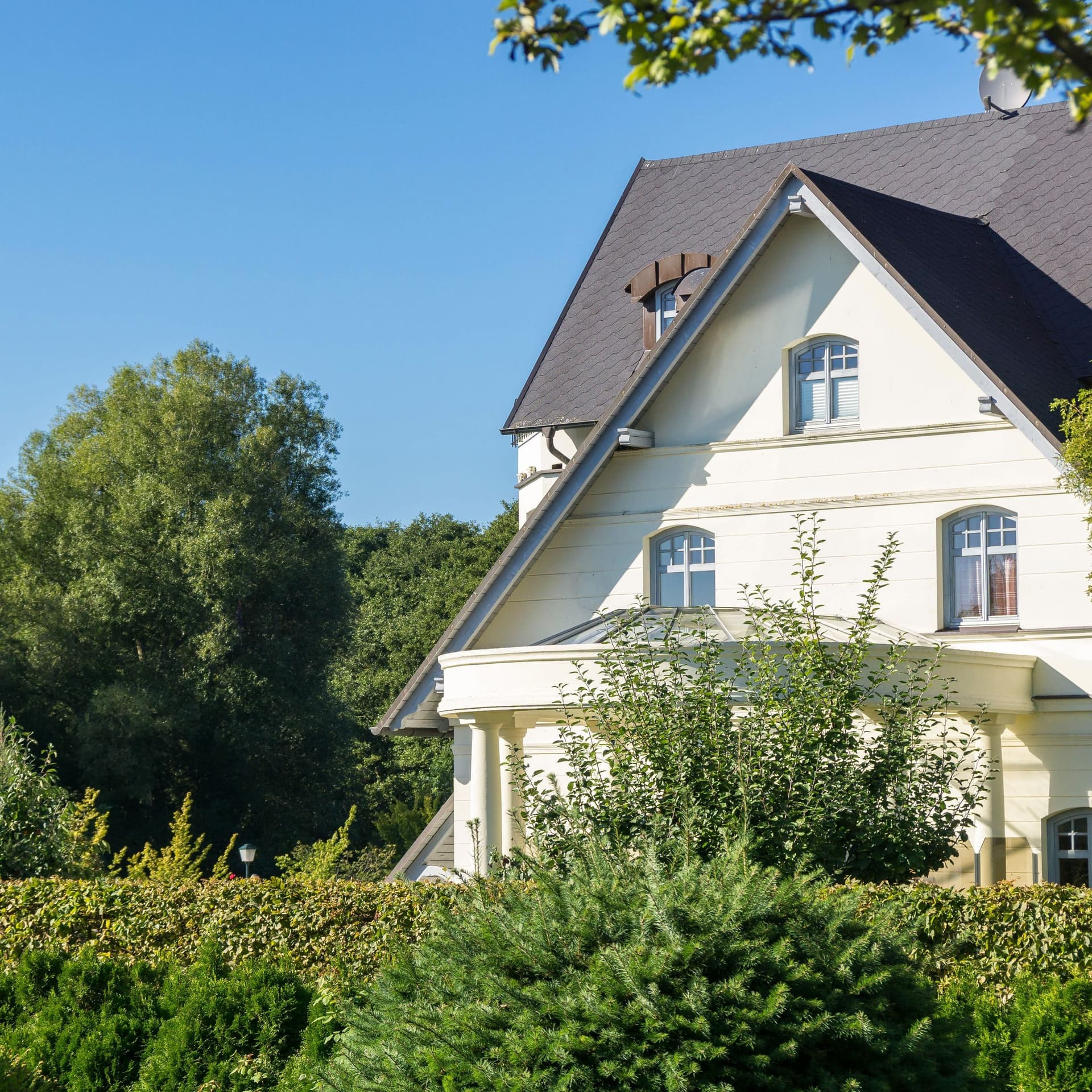 Ein großes weißes Einfamilienhaus hinter Hecke. Das Haus isr umgeben von viel Grün.