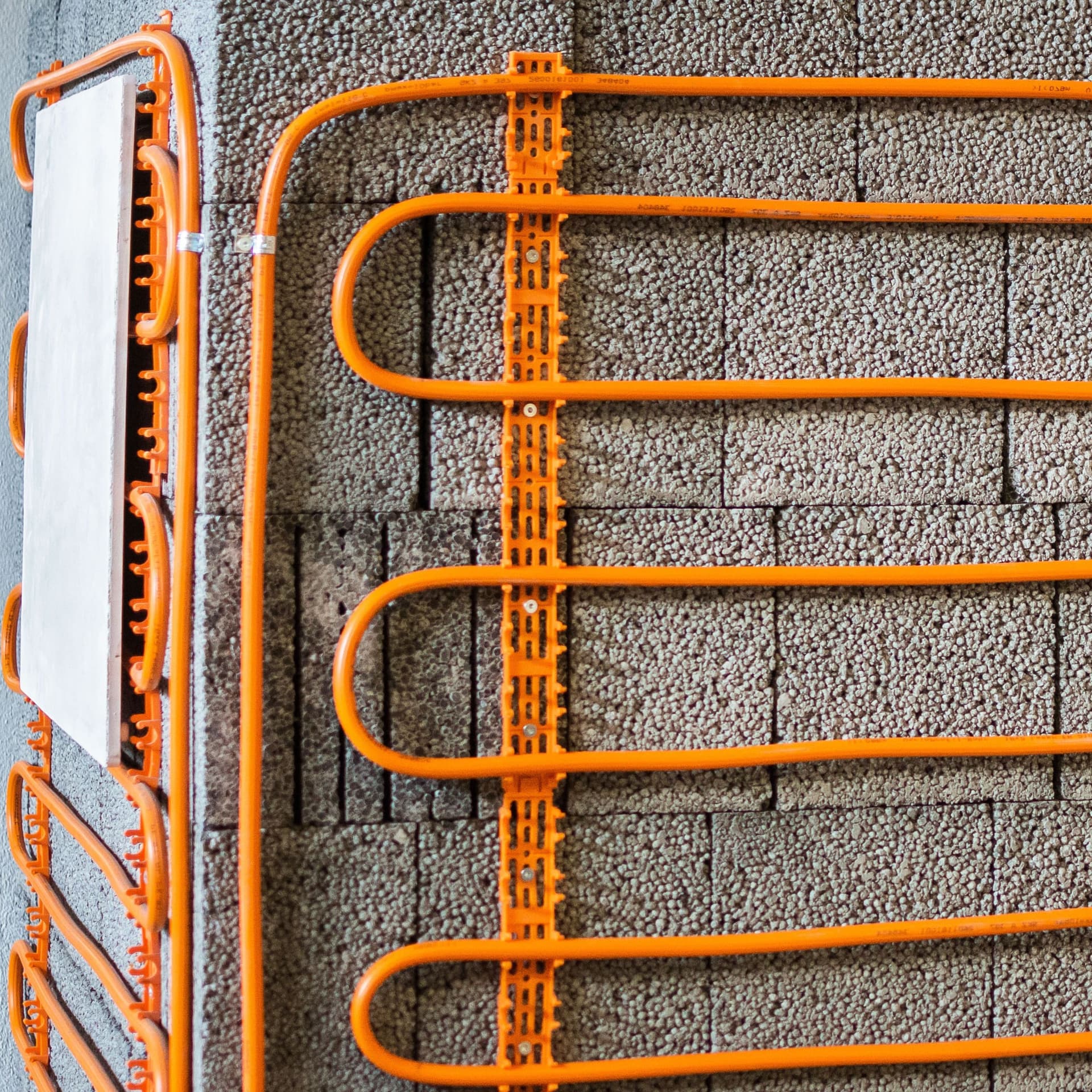 Orangefarbene Kunstoffrohrleitungen für eine Wandheizung mit Halterungen vor unverputzter grauer Mauerwerkswand.