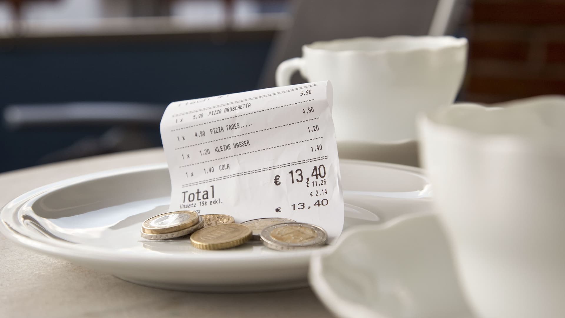 Zwei weiße Kaffeetassen stehen auf einem Restauranttisch. Trinkgeld liegt mit Rechnung auf dem Teller.