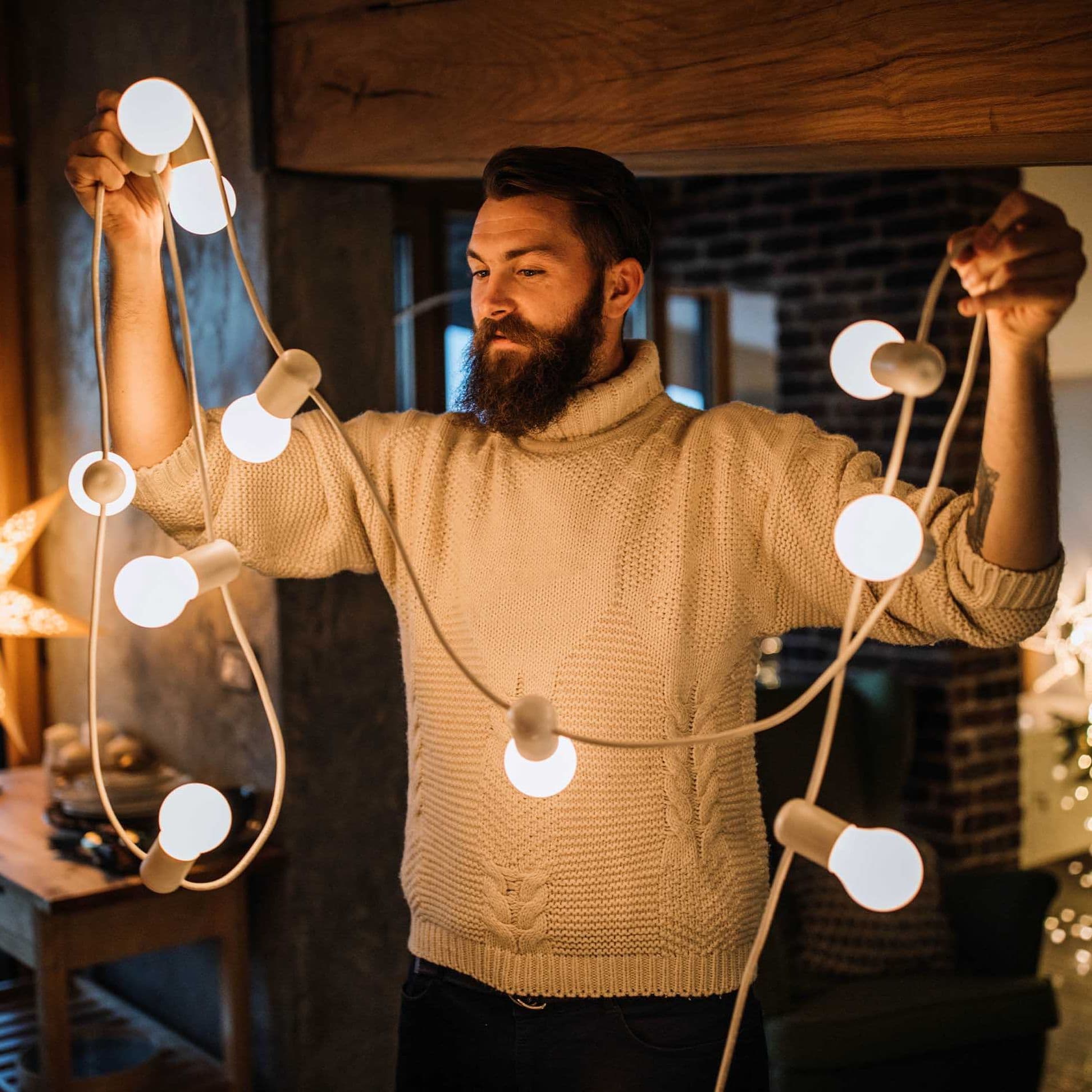 Ein Mann mit Vollbart und hellem Pullover steht im Wohnzimmer. Er hält eine Lichterkette hoch.
