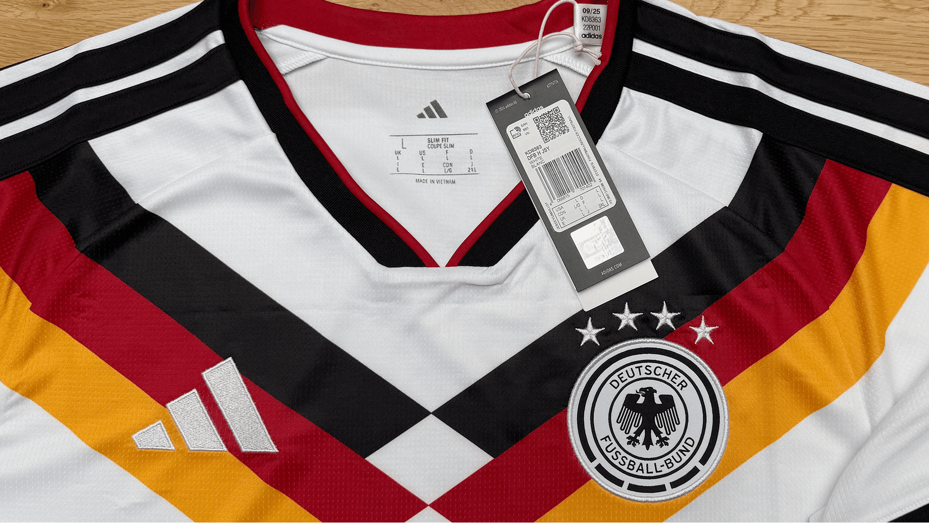 Fußballtrikot in Weiß mit schwarzen, roten und goldenen Streifen, DFB-Logo und Etikett liegt auf einer Holzfläche