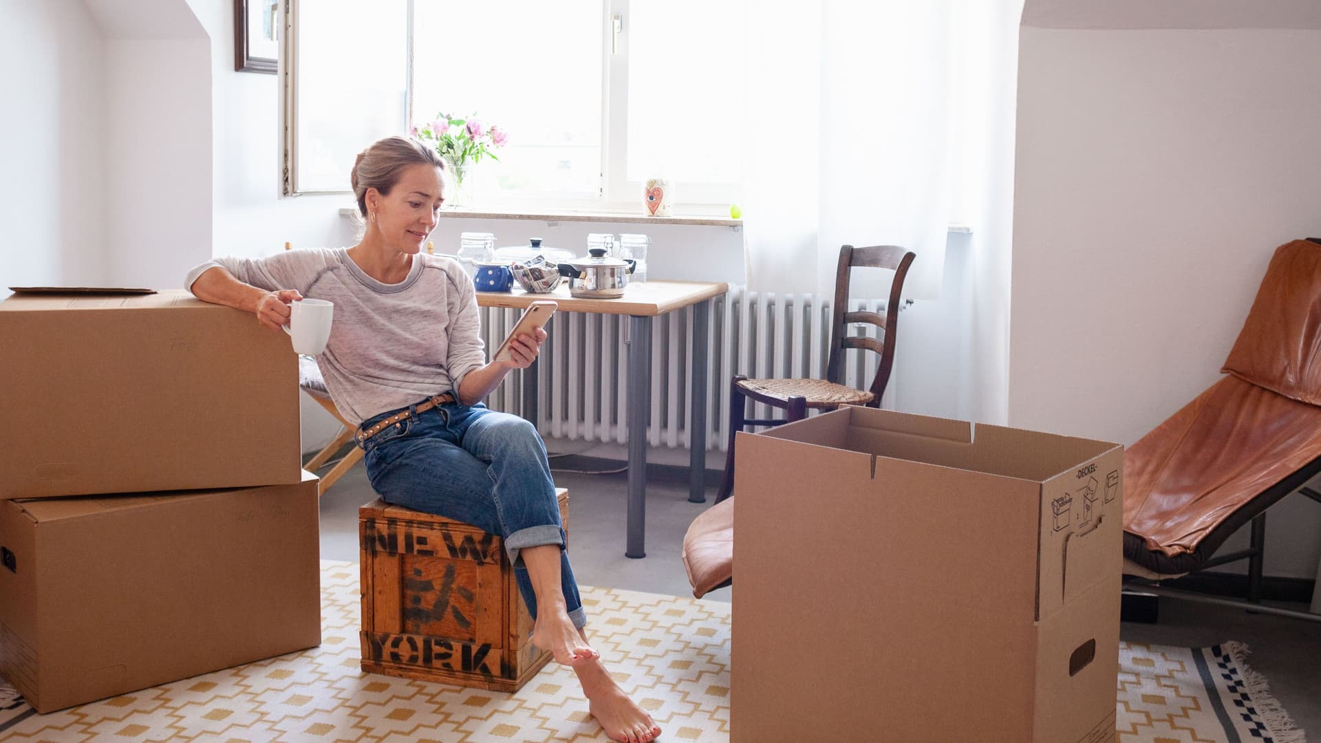 Frau sitzt auf gepackten Kisten in ihrer Wohnung. Sie schaut auf ihr Smartphone.