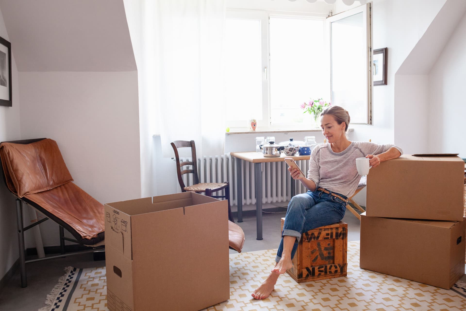 Frau sitzt auf gepackten Kisten in ihrer Wohnung. Sie schaut auf ihr Smartphone.