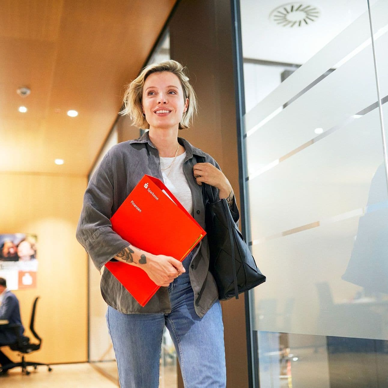 Eine junge Frau in einer Sparkassenfinale. Sie läuft lächelnd mit einer roten Mappe unter dem Arm an einer Glaswand vorbei.