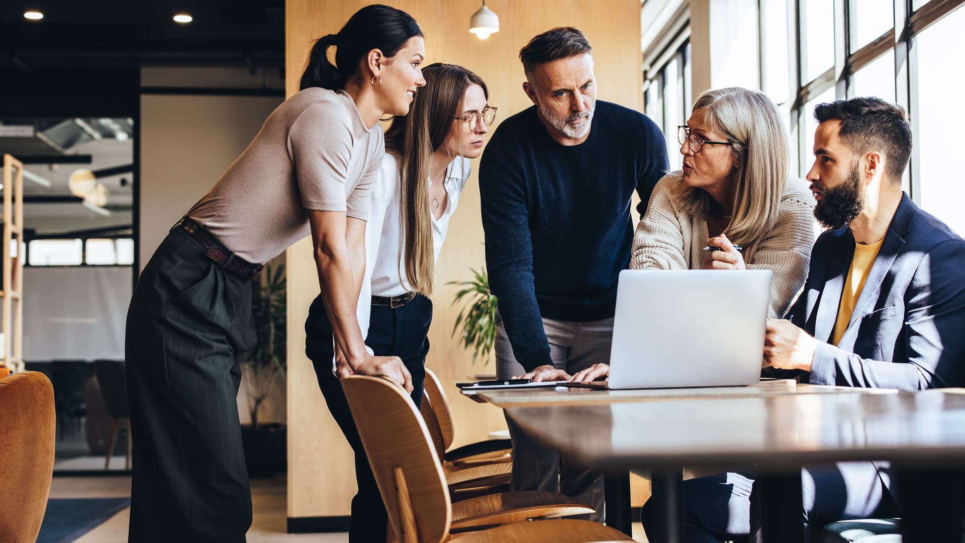 Gruppe aus zwei Männern und drei Frauen steht und sitzt vor einem Laptop an einem Tisch in einem modernen Büro und diskutiert.


