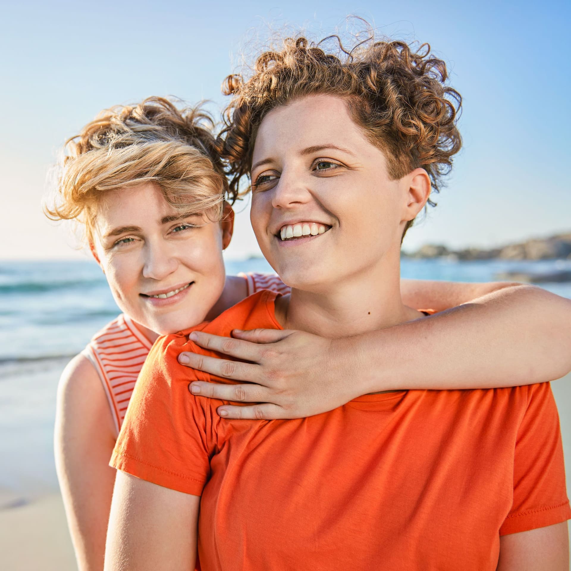 Zwei junge Frauen umarmen sich am Strand. Sie lächeln beide in die Kamera.