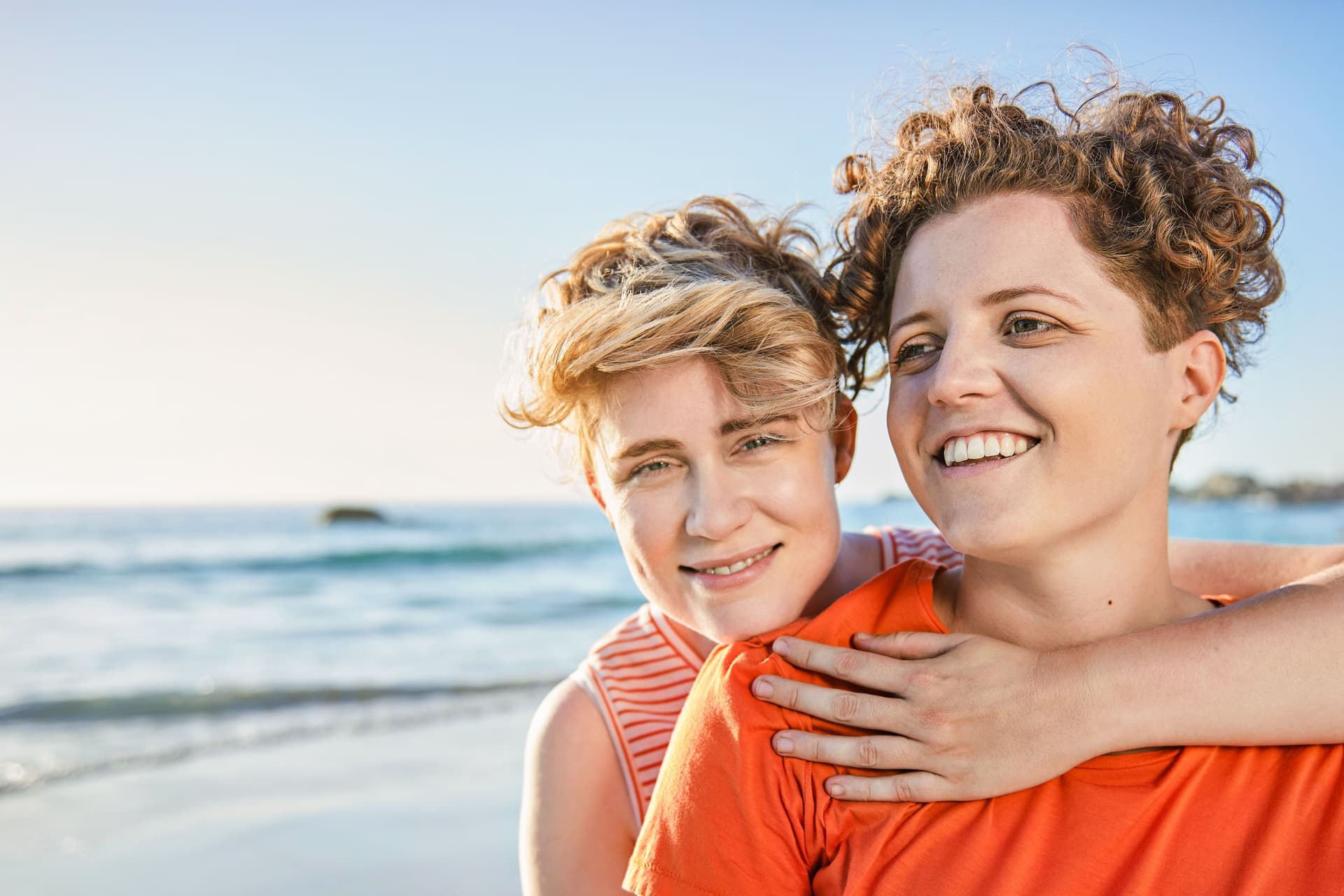 Zwei junge Frauen umarmen sich am Strand. Sie lächeln beide in die Kamera.