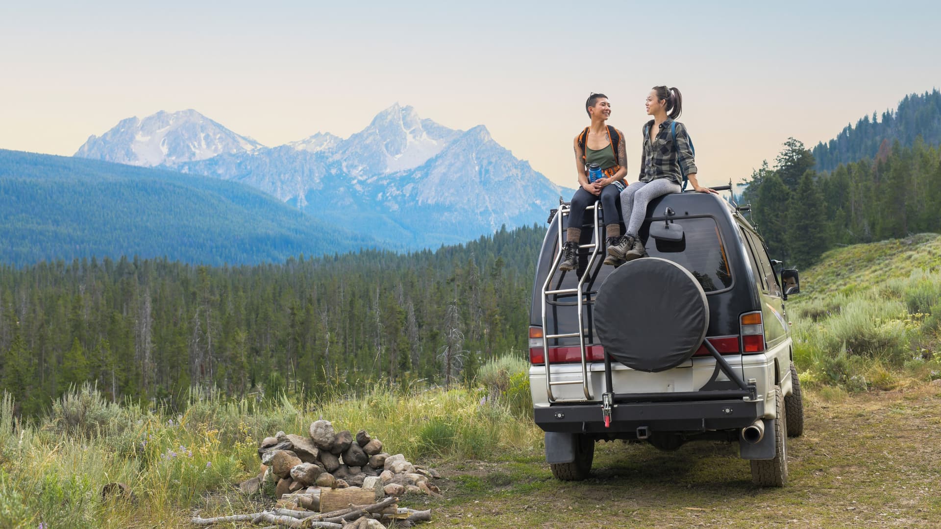 Zwei junge Frauen sitzen auf einem Van. Im Hintergrund Berge.