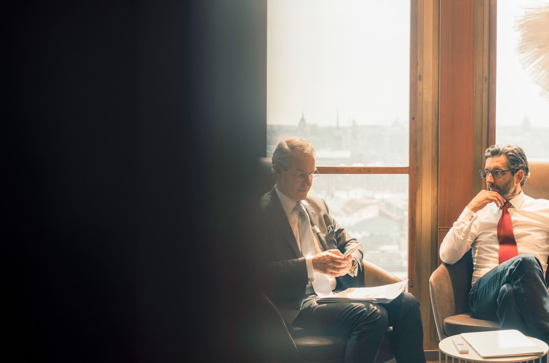 Zwei Männer in Business Kleidung sitzen diskutierend vor einem großen Fenster.