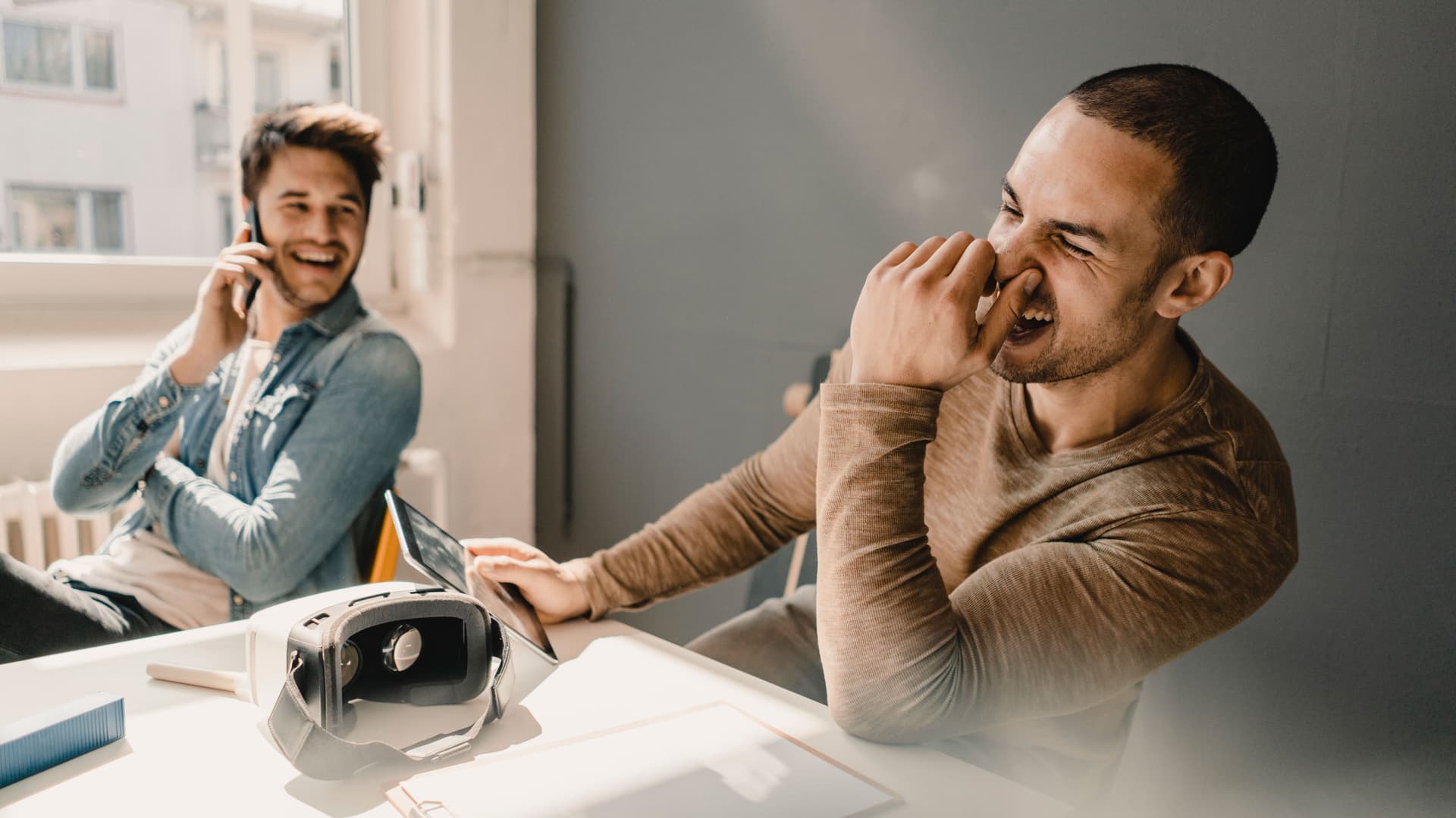 Zwei Männer in einem Büro sitzen nebeneinander und lachen. Einer hat ein Smartphone am Ohr der andere hält sich die Nase zu. Vor ihnen liegt eine Virtuel Reality Brille