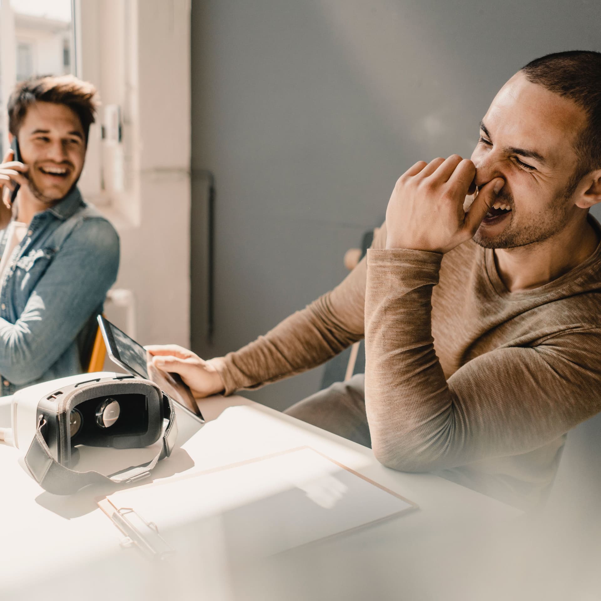 Zwei Männer in einem Büro sitzen nebeneinander und lachen. Einer hat ein Smartphone am Ohr der andere hält sich die Nase zu. Vor ihnen liegt eine Virtuel Reality Brille