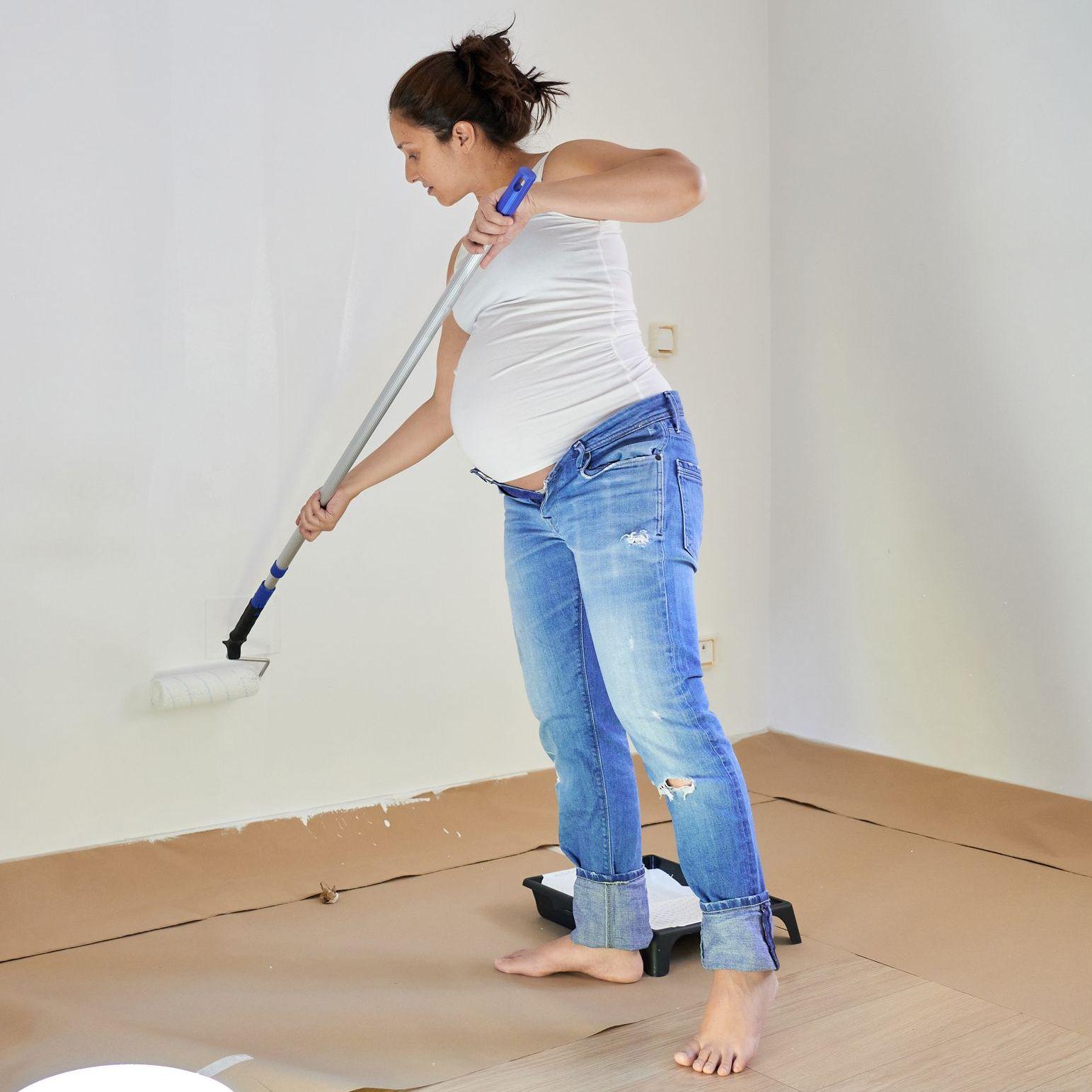 Eine schwangere Frau in Jeans und weißem Shirt malert eine Wand mit weißer Wandfarbe. 