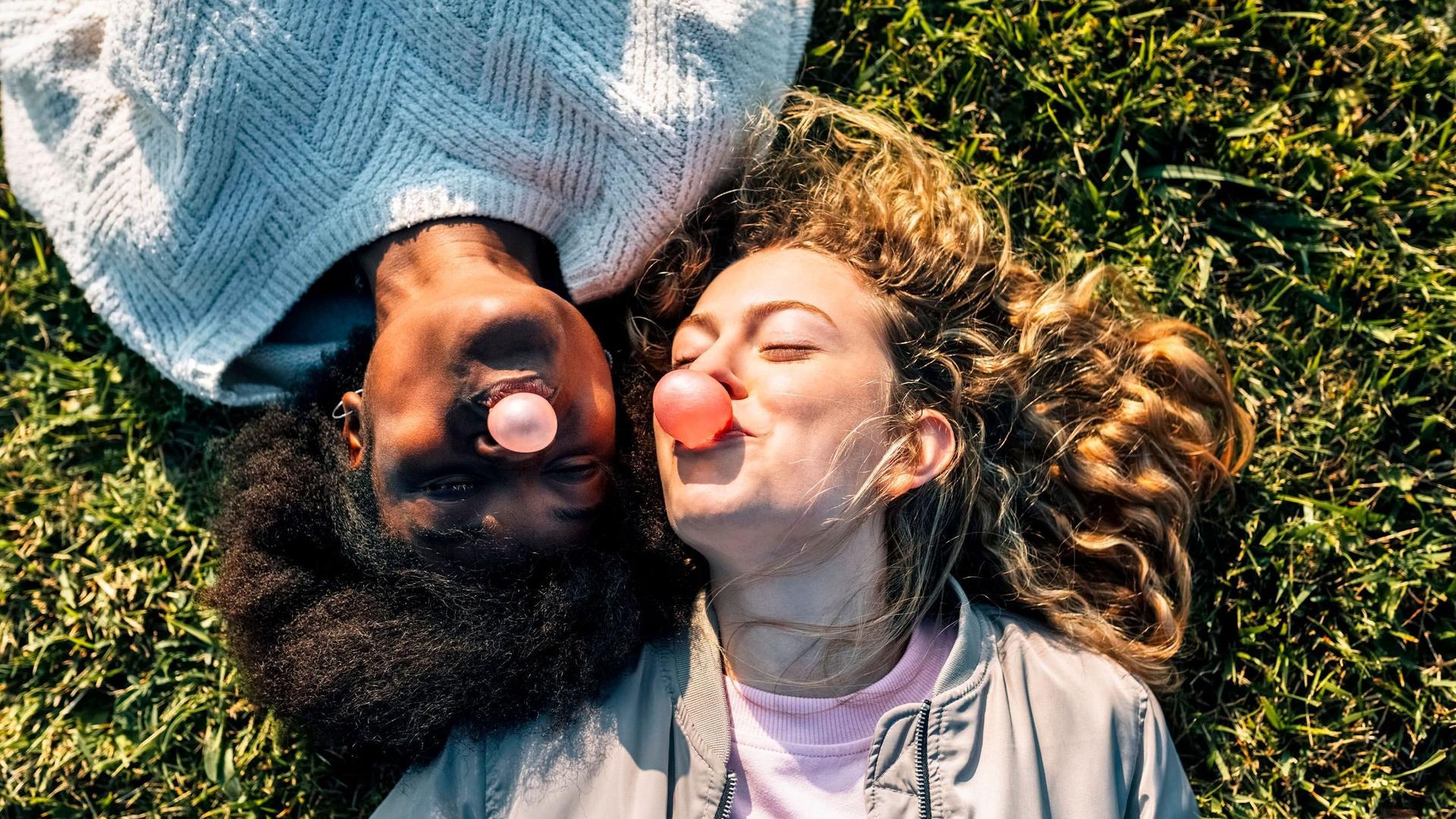 Zwei junge Frauen liegen auf einer grünen  Wiese im Sonnenschein und machen lachend Kaugummiblasen.