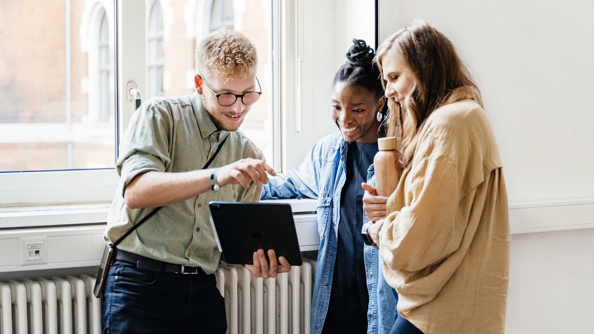 Drei Studenten unterschiedlicher Herkunft stehen zusammen, schauen auf ein Tablet und lächeln.