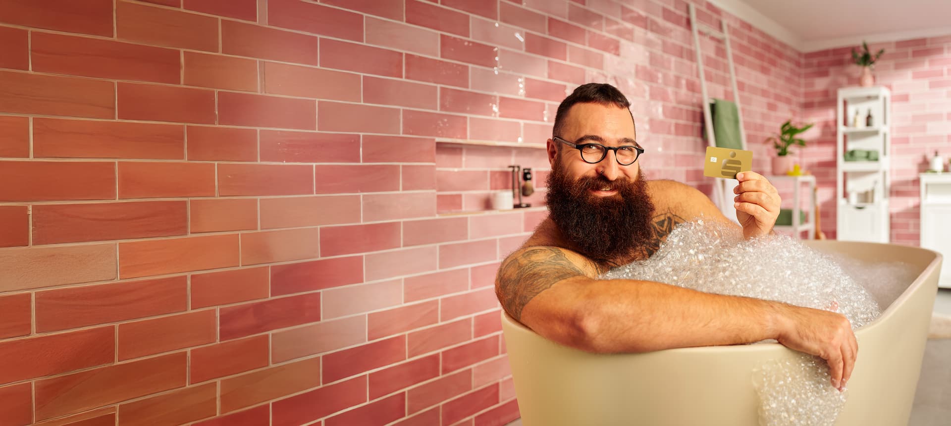 Mann mit Bart und Brille liegt entspannt in einer Badewanne mit Schaum und hält eine goldene Sparkasse-Kreditkarte in die Höhe. Im Hintergrund eine rose-farbene Fliesenwand.