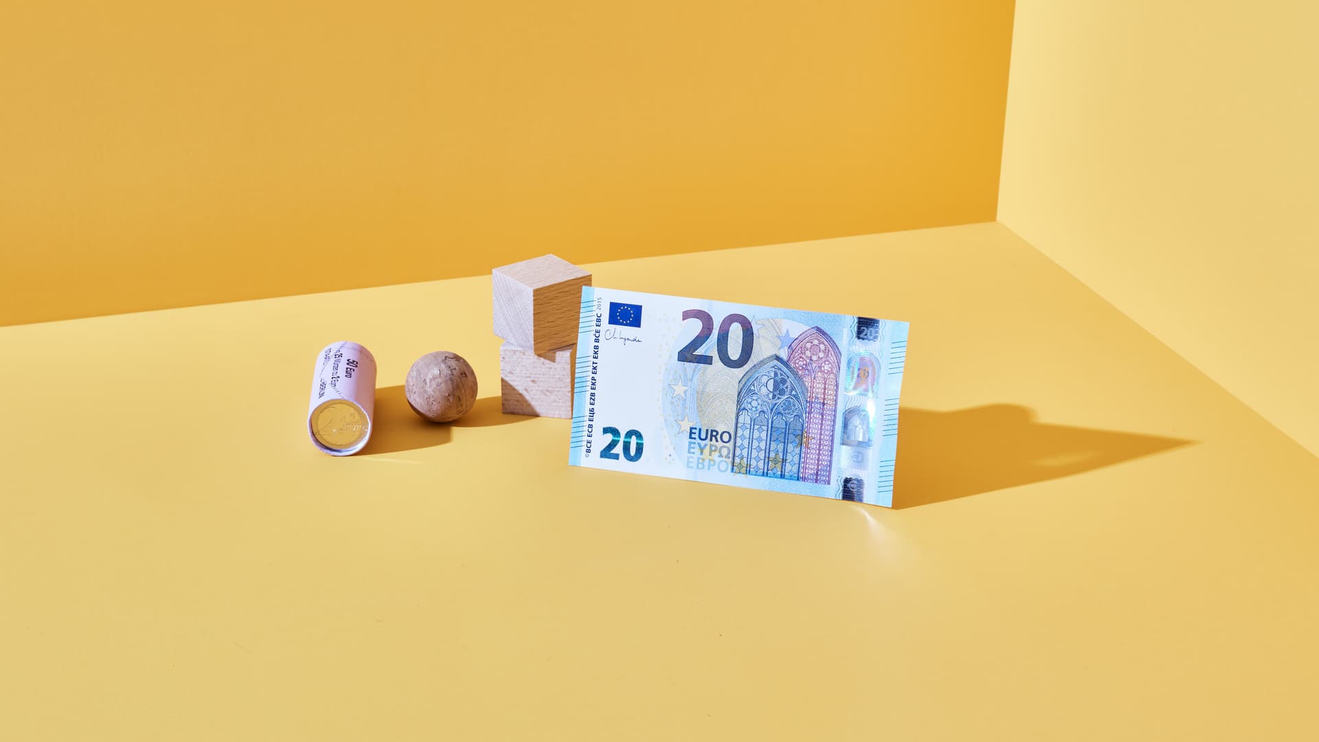 20-Euro-Schein steht aufrecht neben Holzwürfel, Murmel und Münzrolle auf gelbem Untergrund