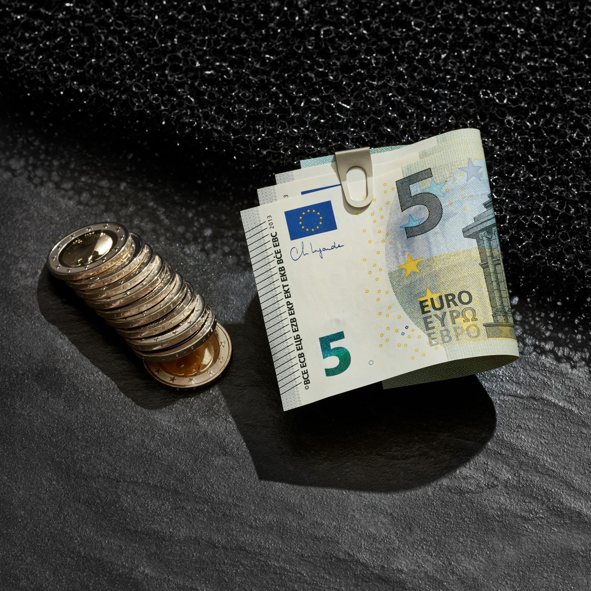 Münzstapel und gefaltete 5-Euro-Scheine mit Büroklammer auf dunklem Untergrund
