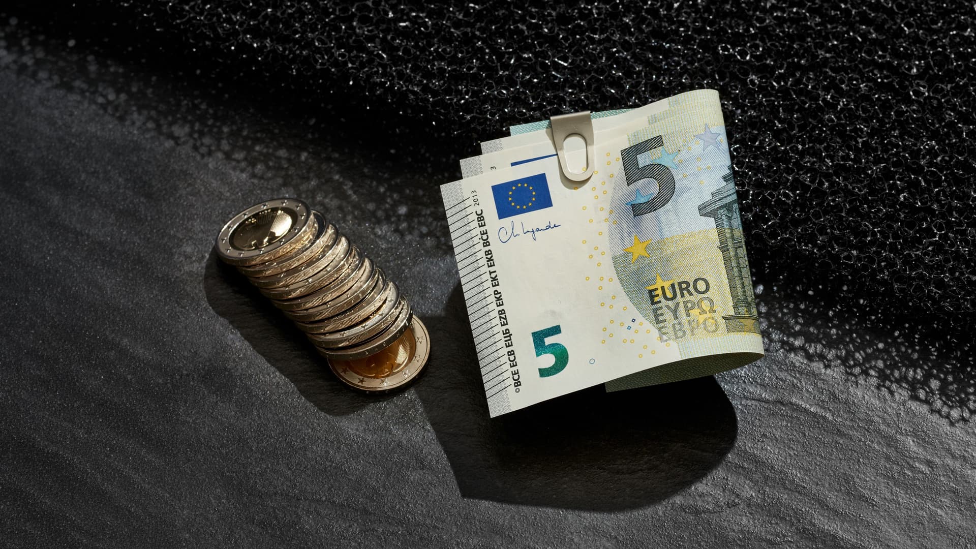 Münzstapel und gefaltete 5-Euro-Scheine mit Büroklammer auf dunklem Untergrund