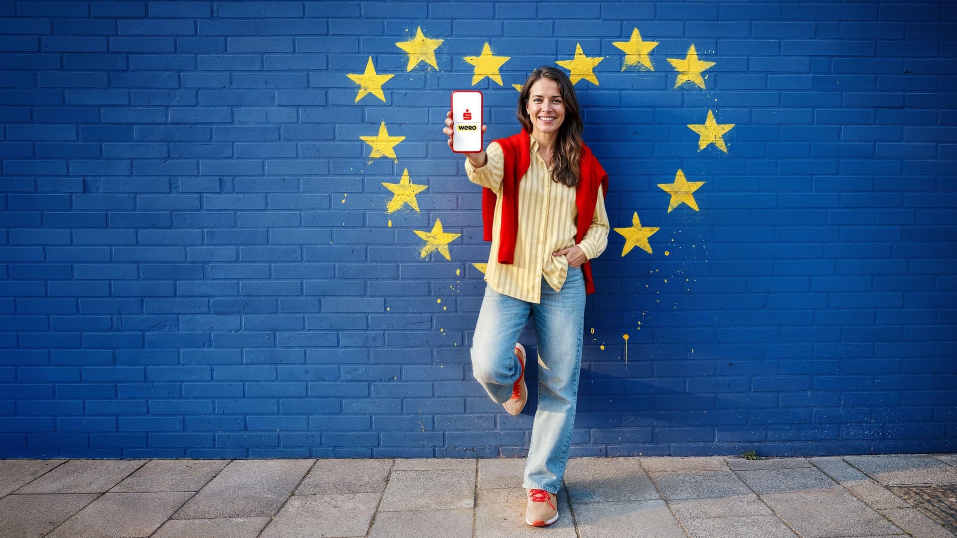 Eine lächelnde Frau steht vor einer blauen Backsteinmauer, die mit den Sternen der Europäischen Union bemalt ist. Sie hält ein Smartphone mit dem "wero"-App-Logo hoch.