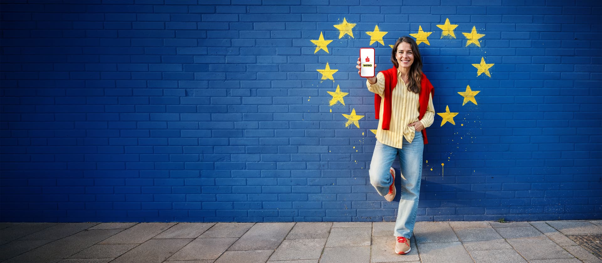 Eine lächelnde Frau steht vor einer blauen Backsteinmauer, die mit den Sternen der Europäischen Union bemalt ist. Sie hält ein Smartphone mit dem "wero"-App-Logo hoch.