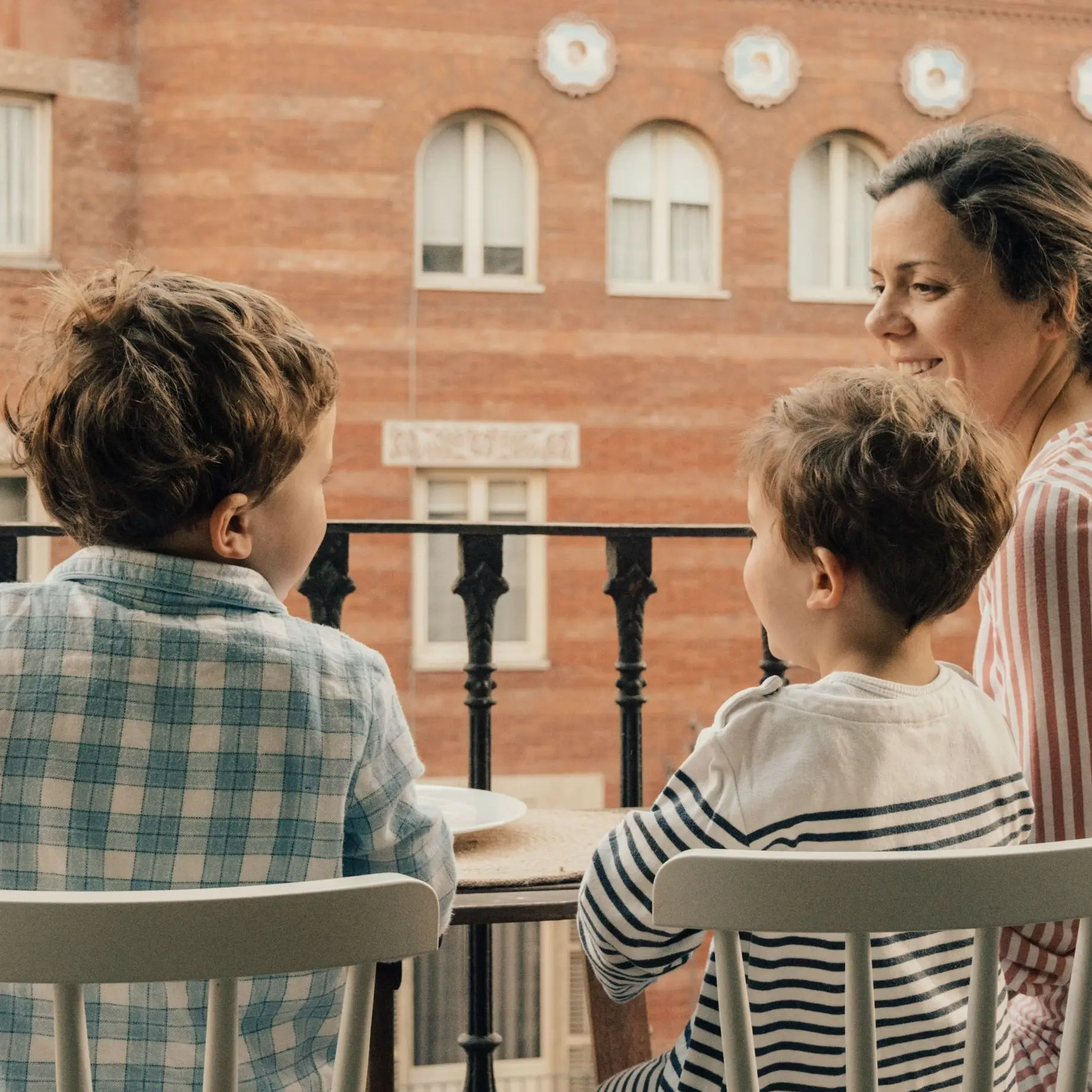 Eine Frau sitzt mit zwei Kindern auf einem Balkon in einer städtischen Umgebung. Im Hintergrund sind Wohnhäuser zu sehen.
