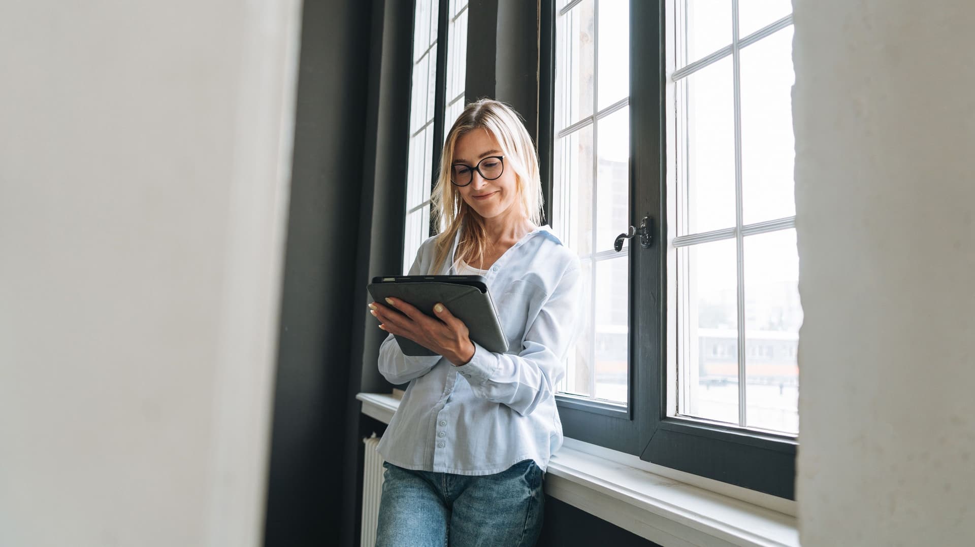 Eine junge Frau lehnt an einem Fenster und blickt auf ihr Tablet