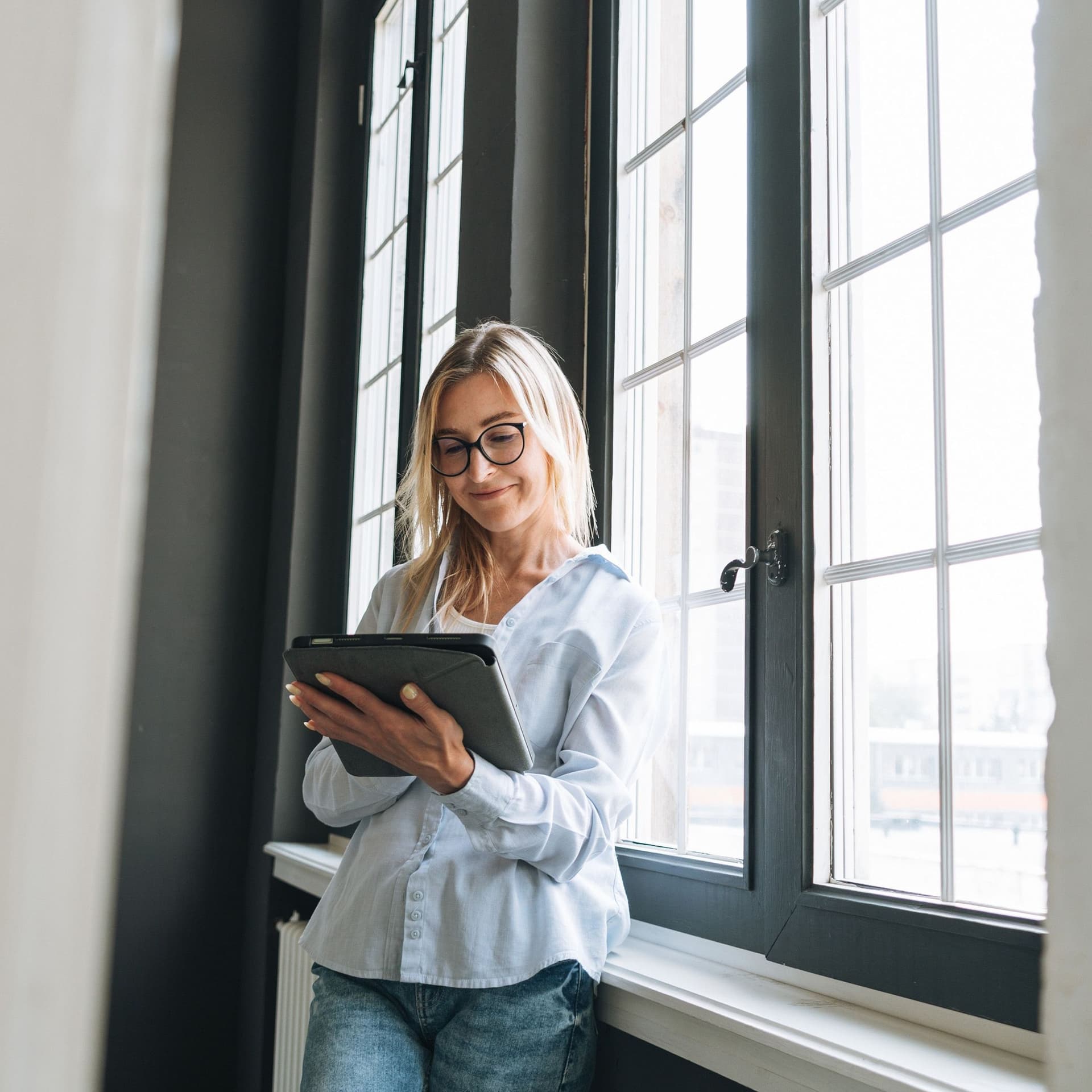 Eine junge Frau lehnt an einem Fenster und blickt auf ihr Tablet