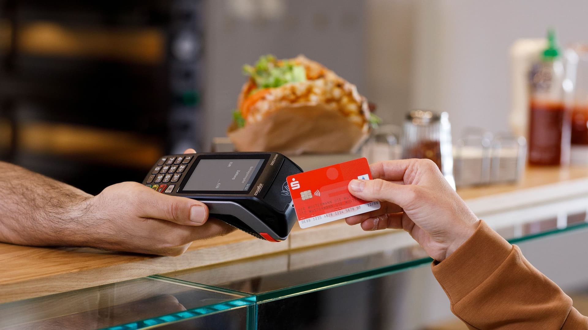 Hand hält rote Sparkassen-Karte an Kartenterminal, Imbiss mit Döner im Hintergrund