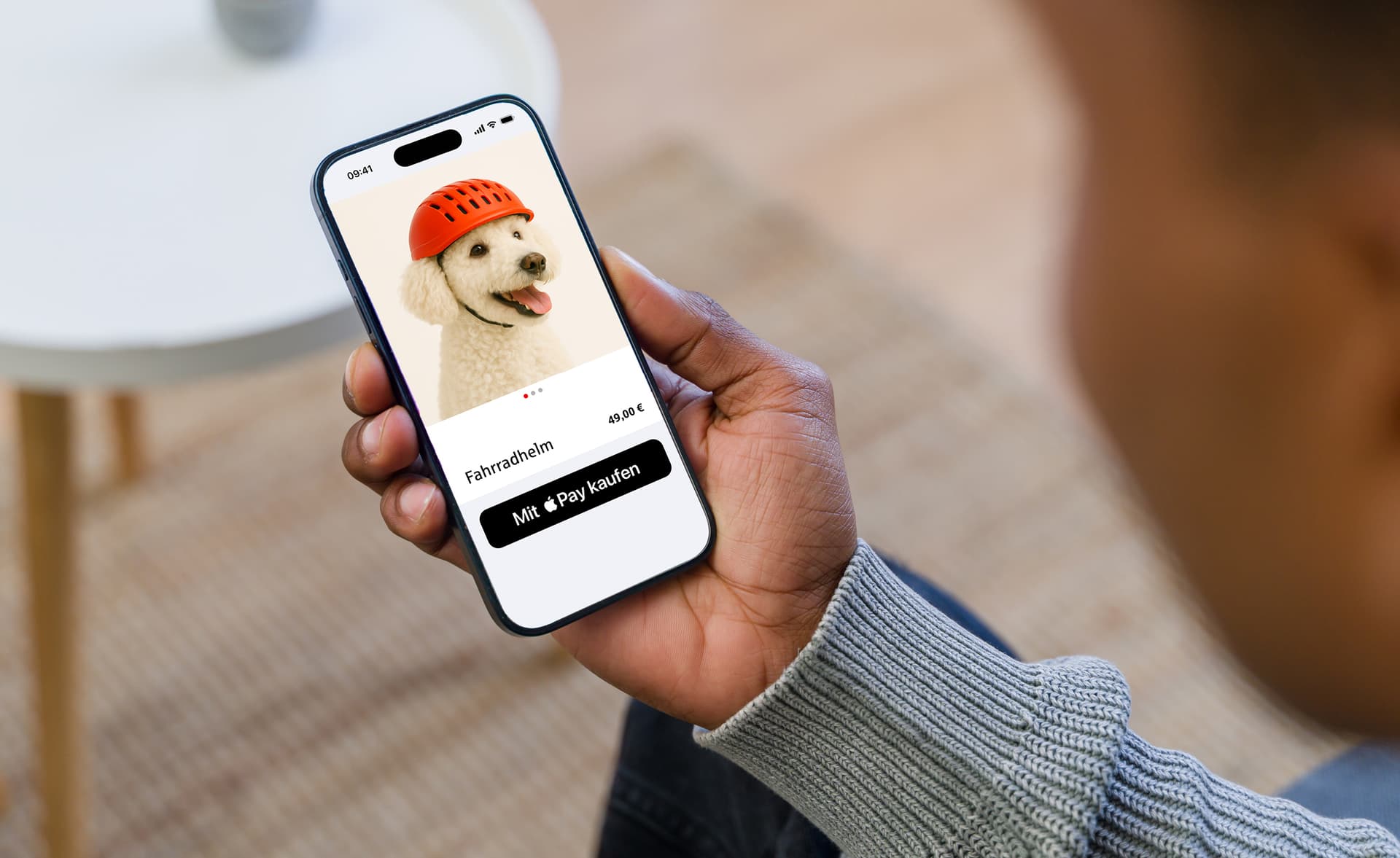 Person hält Smartphone mit einem weißen Pudel, der einen orangefarbenen Fahrradhelm trägt, auf einer E-Commerce-Seite mit der Aufschrift 'Fahrradhelm' und 'Mit Apple Pay kaufen'.