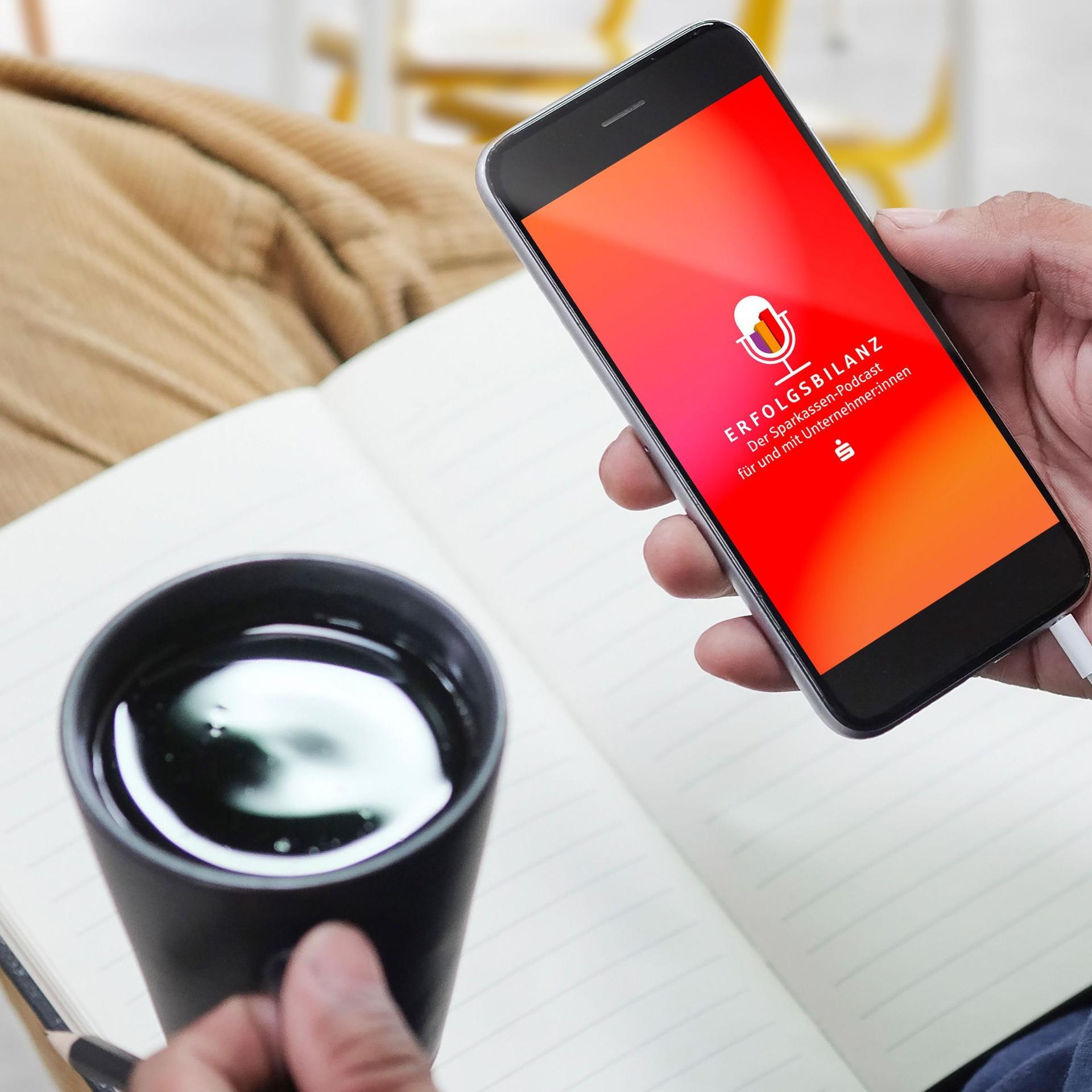 in Notizbuch liegt auf einem Bein.Kaffeetasse in der linken Hand. Smartphone in der rechten Hand. Das Display ist orange mit Logo Erfolgsbilanz.