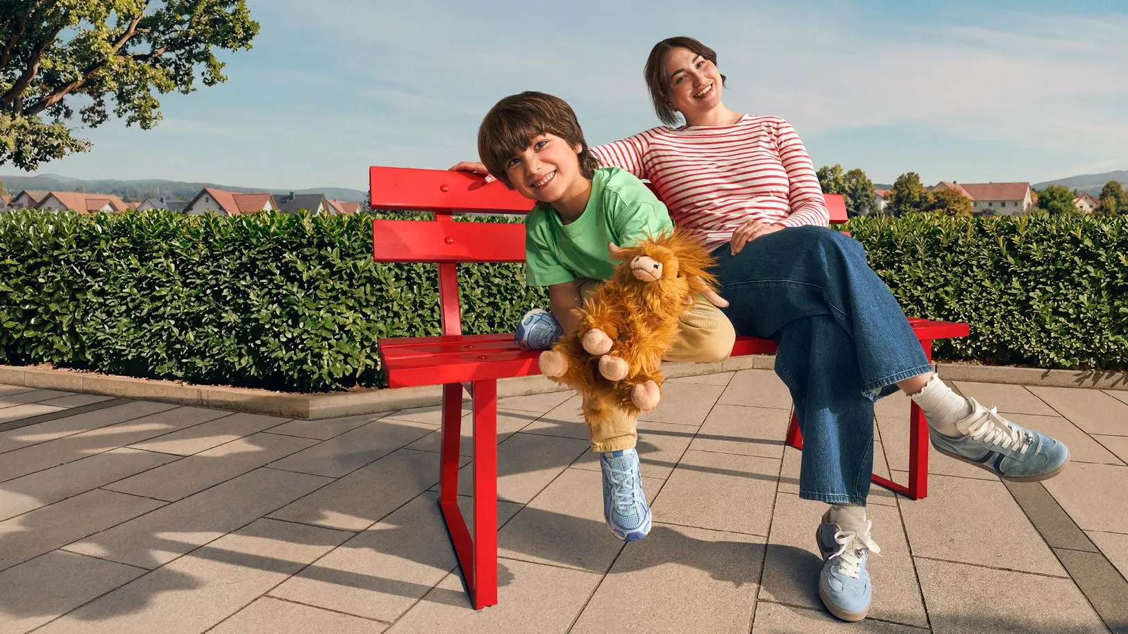 Mutter und Kind sitzen lächelnd auf einer roten Bank im Freien, Kind hält ein Kuscheltier