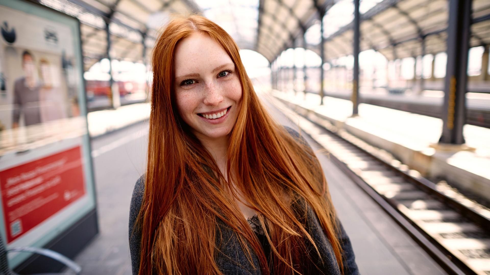 Eine Frau mit langen roten Haaren steht auf einem Bahnsteig.