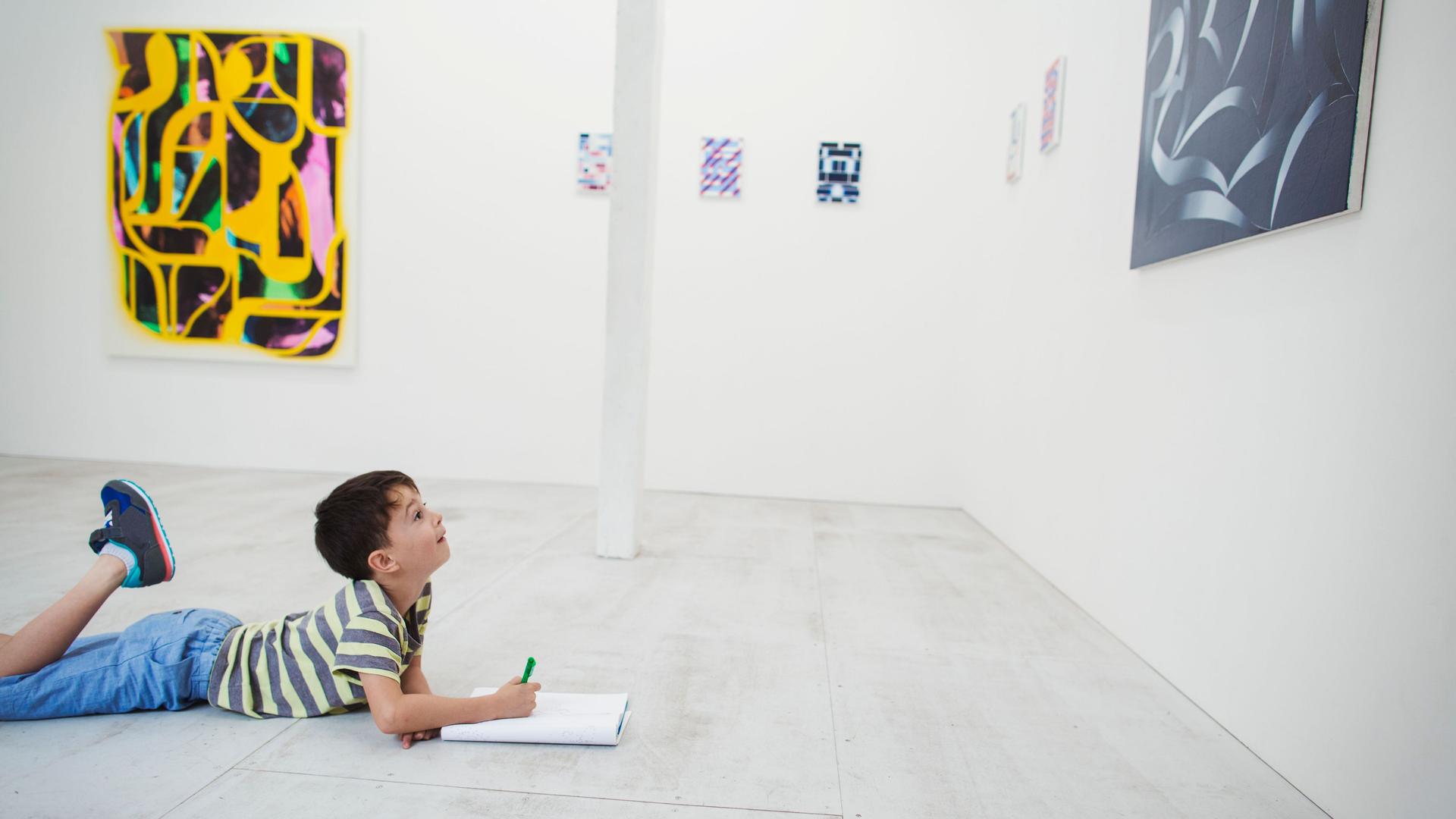 Boy with short black hair lying on floor in art gallery with pen and paper, looking at modern painting.
