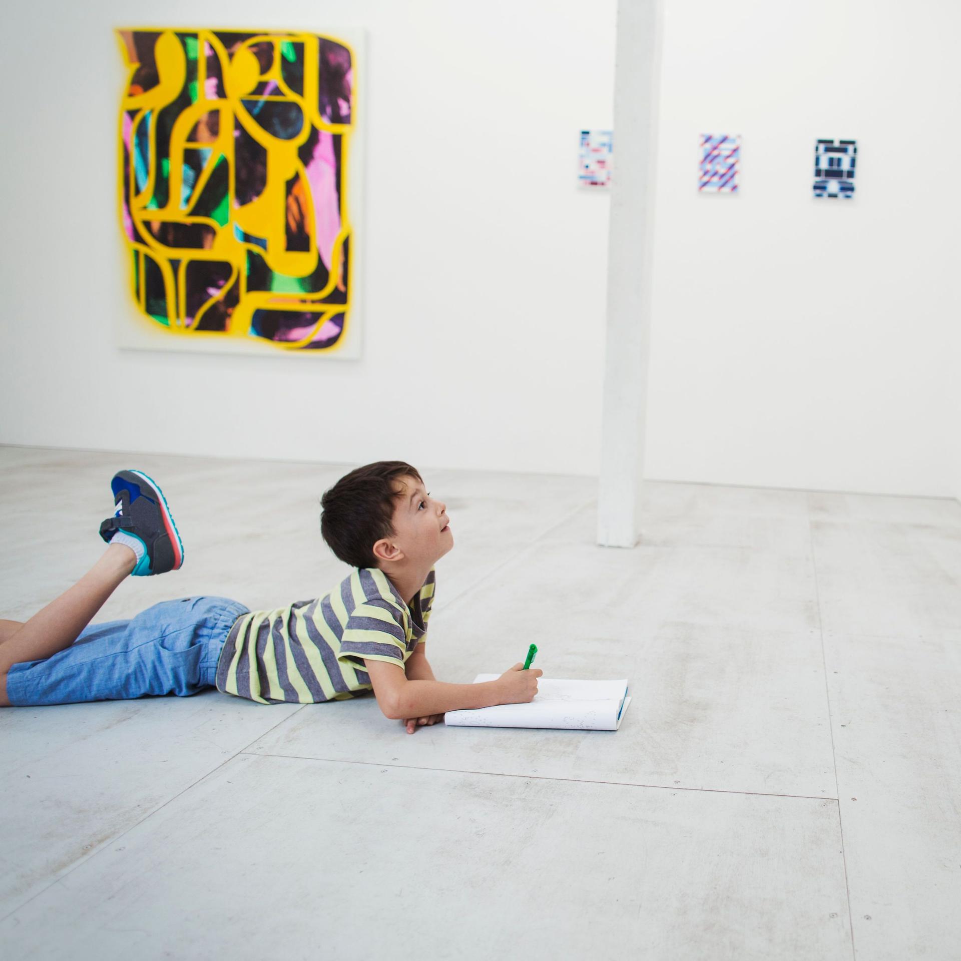 Boy with short black hair lying on floor in art gallery with pen and paper, looking at modern painting.