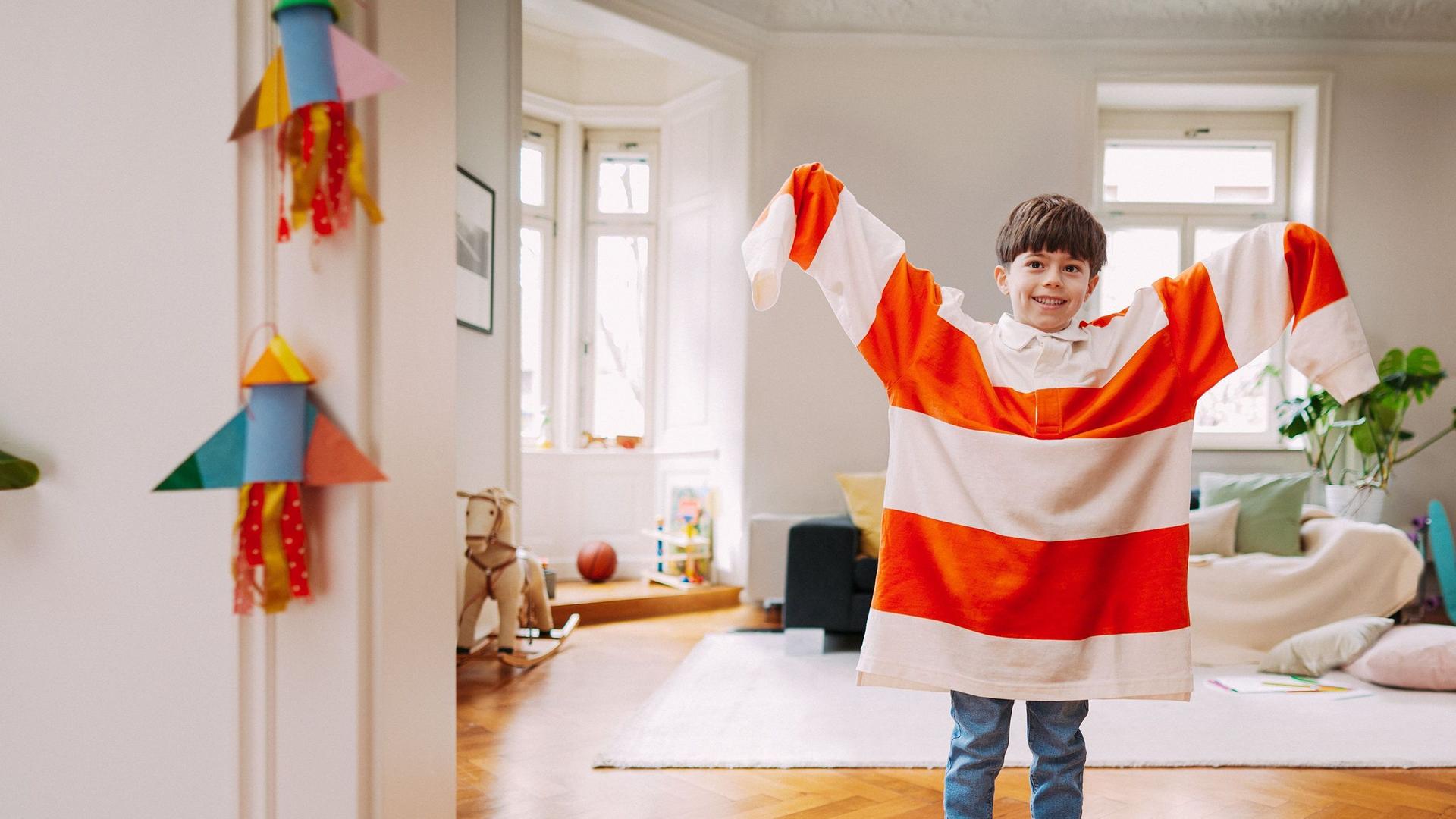 Kleiner Junge steht im mit einem zu großen rot-weiß-gestreiften Pulli im Wohnzimmer und hebt die Arme nach oben