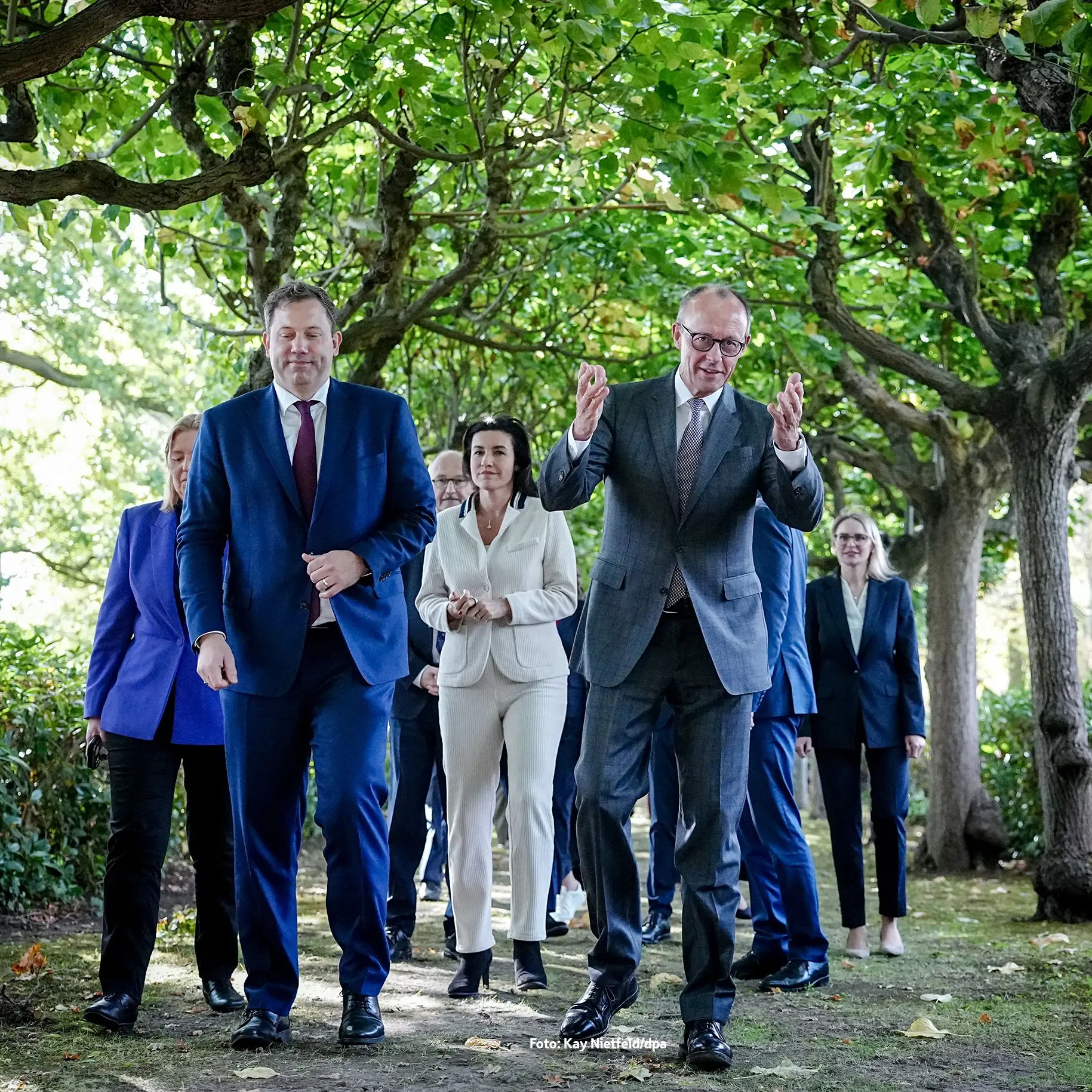 Eine Gruppe von Politikerinnen und Politikern geht im Grünen unter einem Blätterdach entlang. Im Vordergrund spricht Friedrich Merz im grauen Anzug, während Lars Klingbeil im blauen Anzug, Dorothee Bär, Bärbel Bas und weitere Personen folgen.