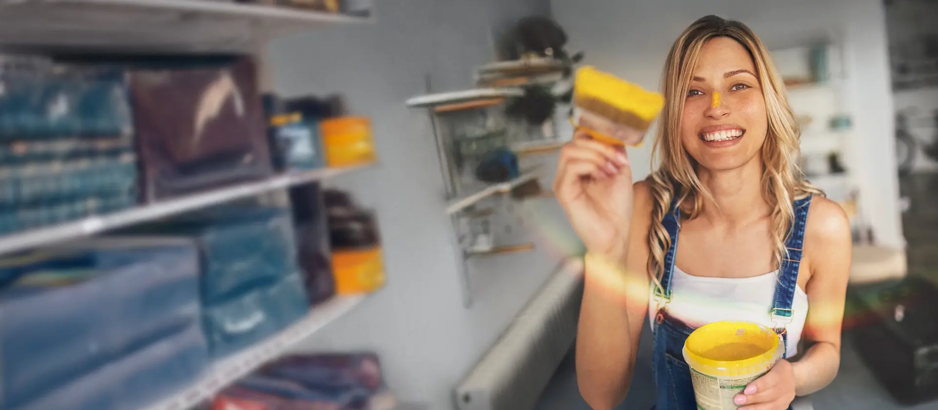Eine lachende junge Frau in Latzhose hält einen gelben Farbtopf und Pinsel in der Hand. Sie steht in einem hellen Raum mit Regalen im Hintergrund und hat gelbe Farbe auf der Nasenspitze – sie wirkt fröhlich.