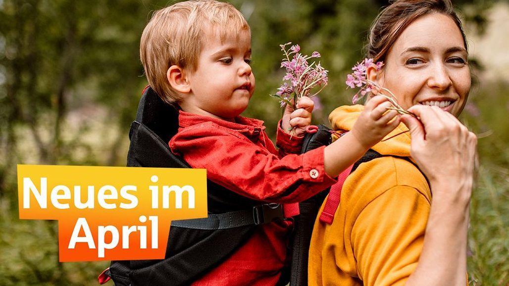 Eine lächelnde Frau im gelben Pulli trägt auf ihrem Rücken einen Jungen, dem sie Blumen recht. Auf dem Bild steht "Neues im April"