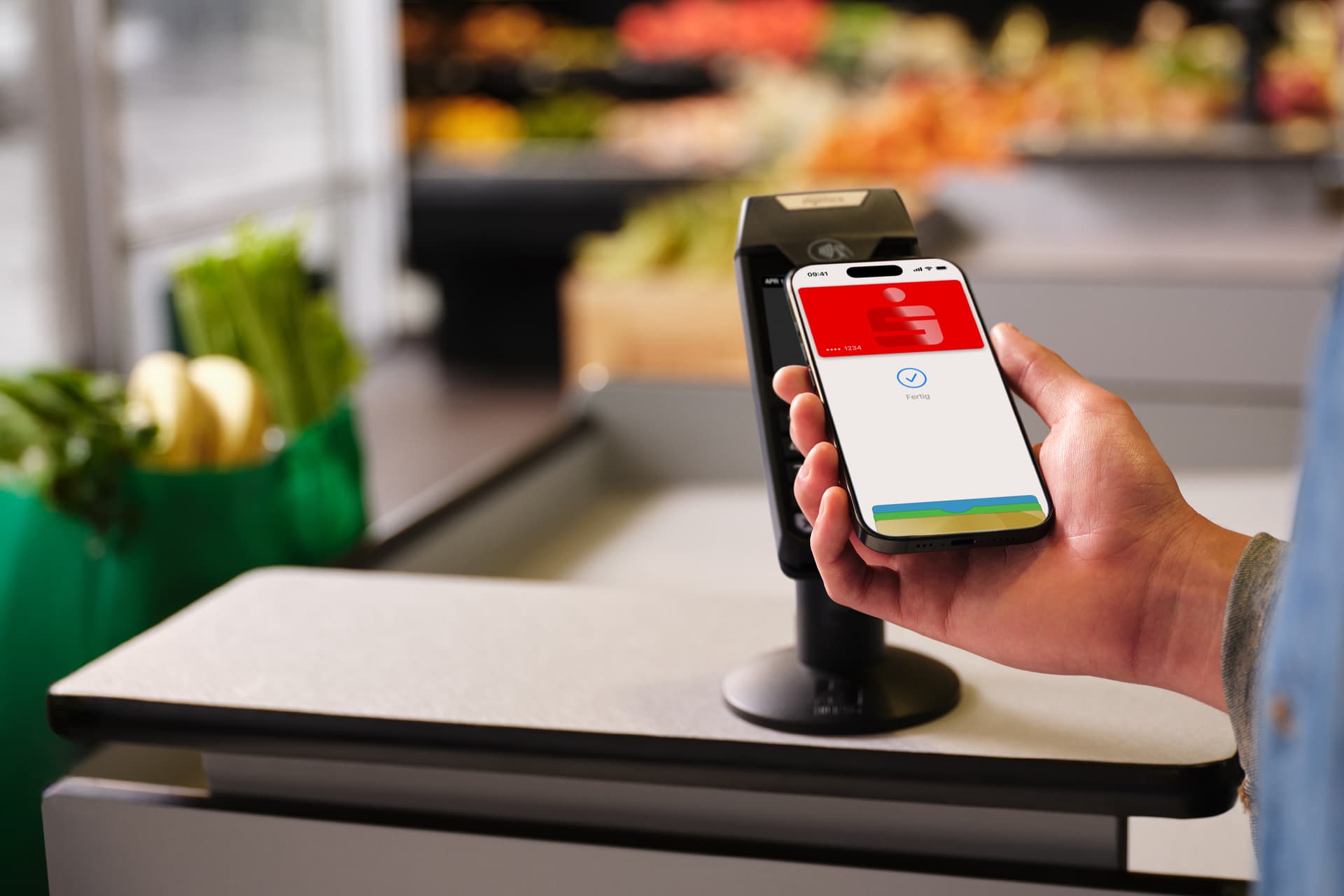 Eine Hand hält ein Smartphone mit einer Sparkassen-Karte in einer Supermarktkasse, bereit für eine kontaktlose Zahlung.