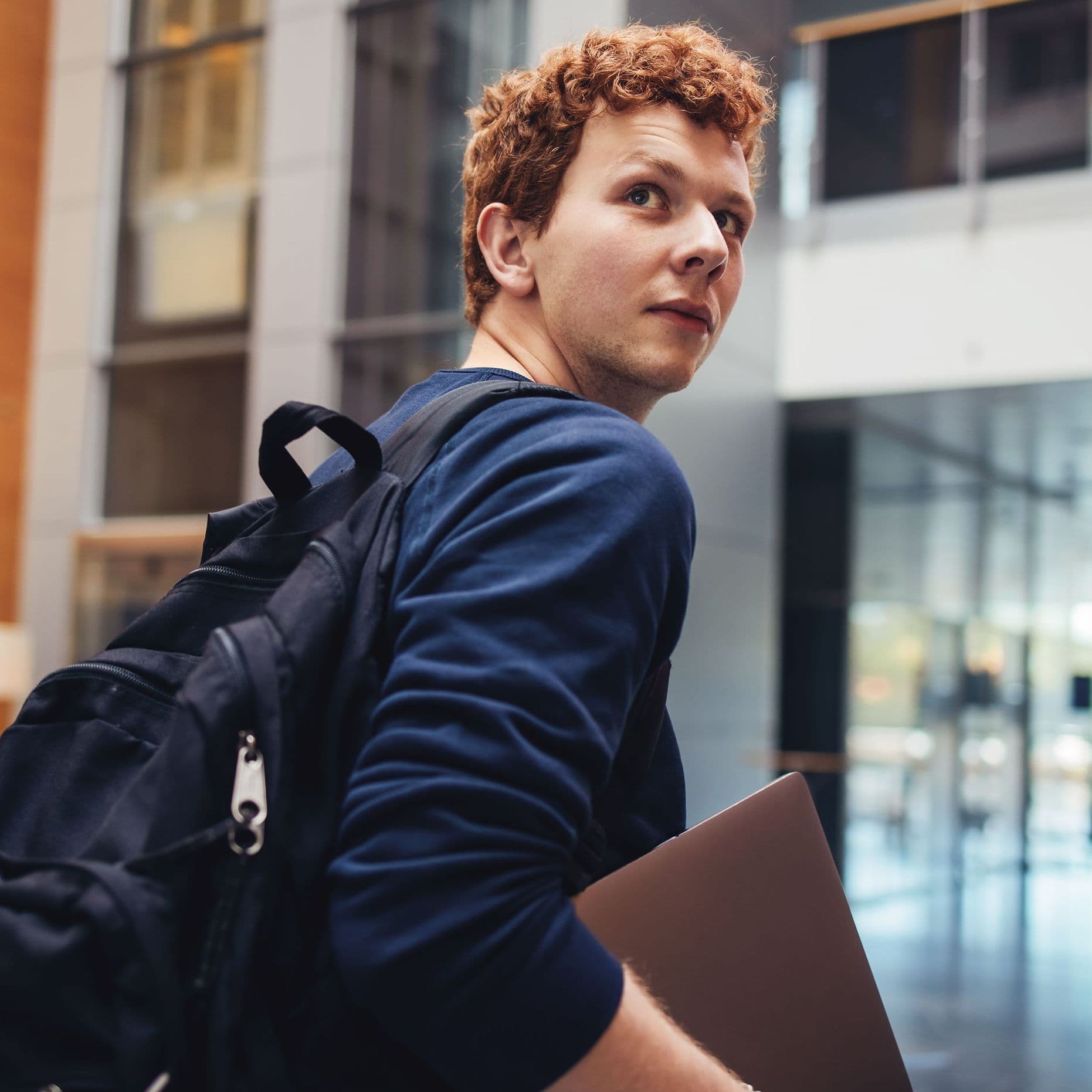 Student mit Laptop unterm Arm blickt über seine Schulter Richtung Kamera.