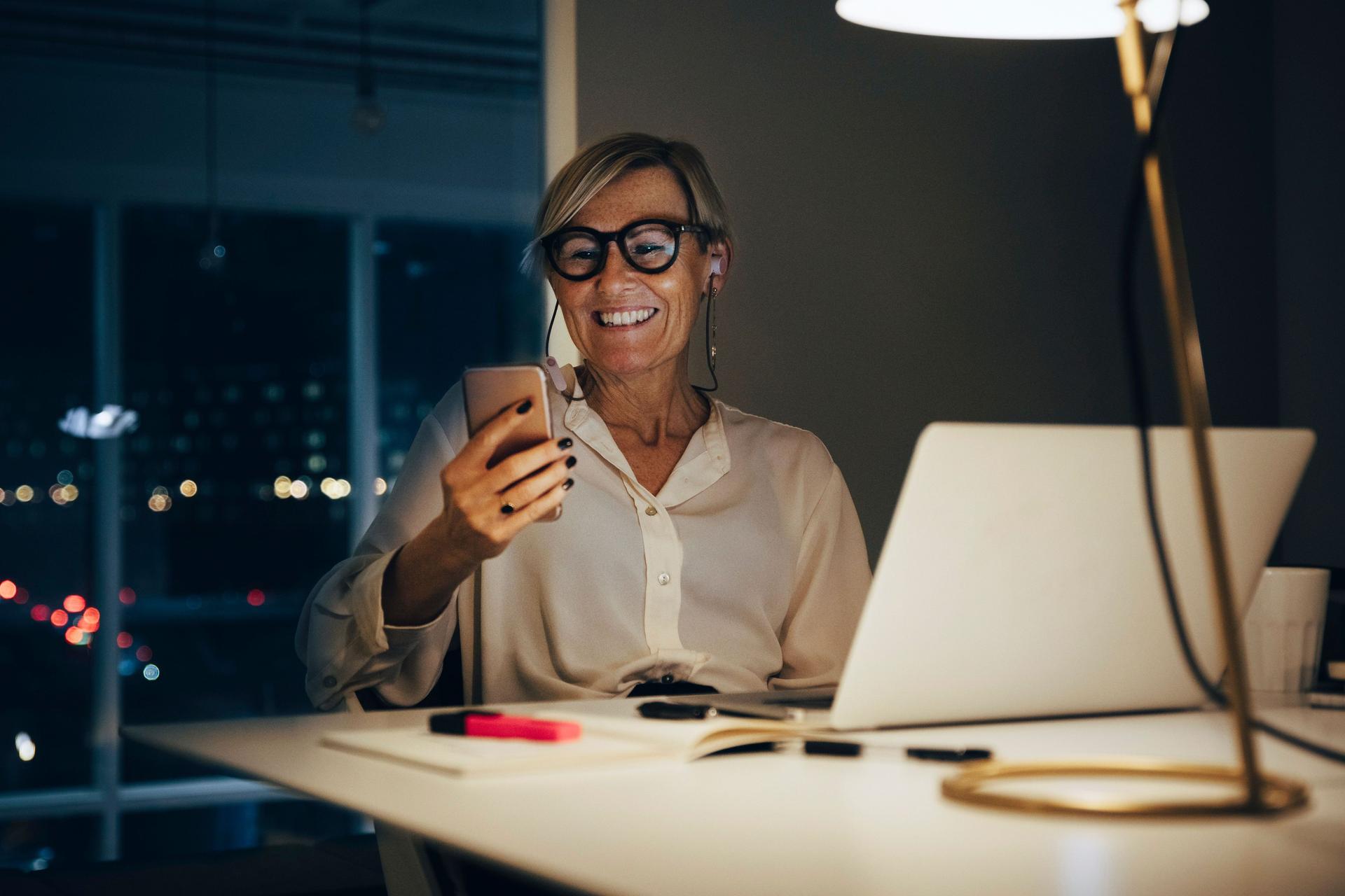 Lächelnde Frau sitzt mit Smartphone in der Hand an einem Schreibtisch. Ihr Laptop steht auf dem Tisch. Durch das Fenster sieht man Lichter einer Stadt, es ist dunkel.