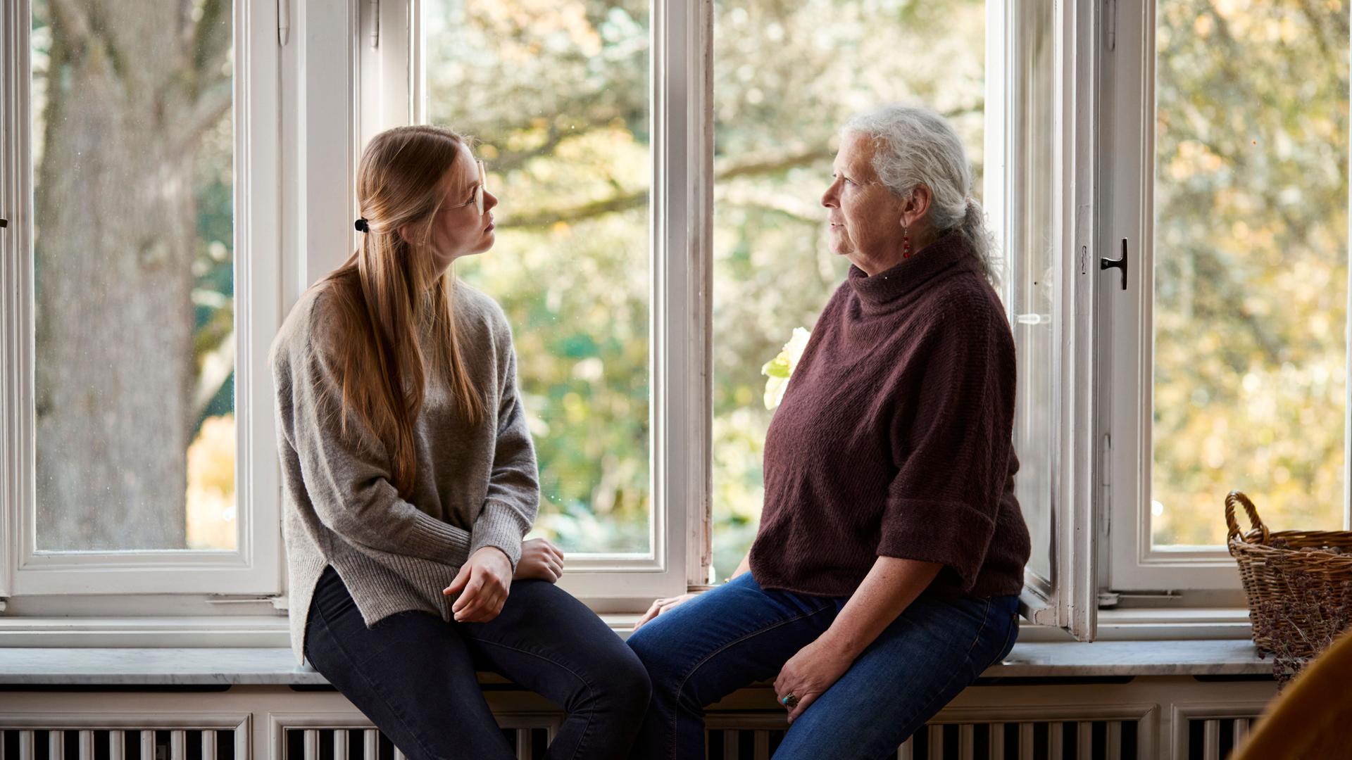 Ältere Frau und junge Frau sitzen sich zugewandt auf der Fensterbank. Hinter ihnen sind Altbaufenster mit Blick ins Grüne.